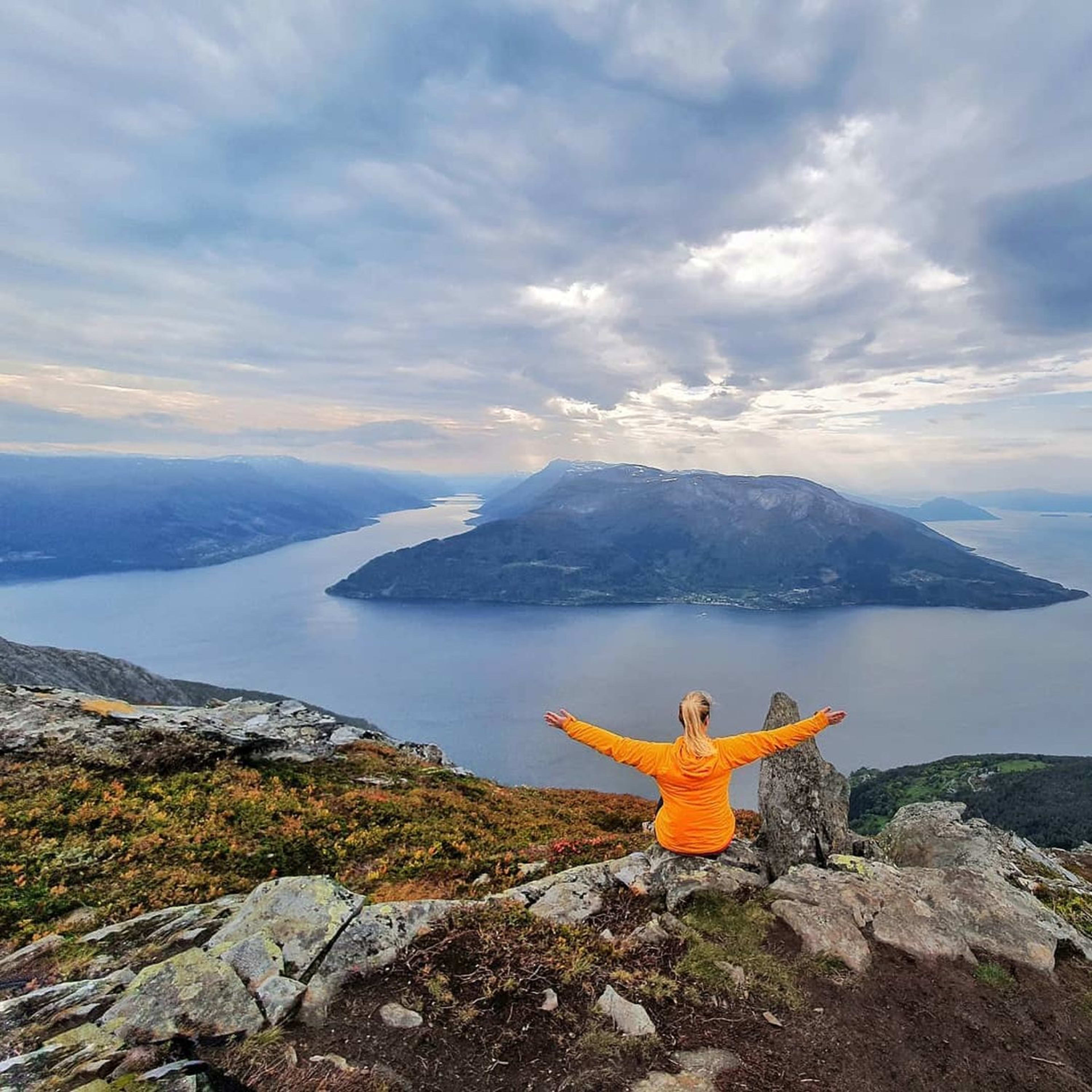 Person in orange sits atop mountain, arms wide, view of the Hardangerfjord.