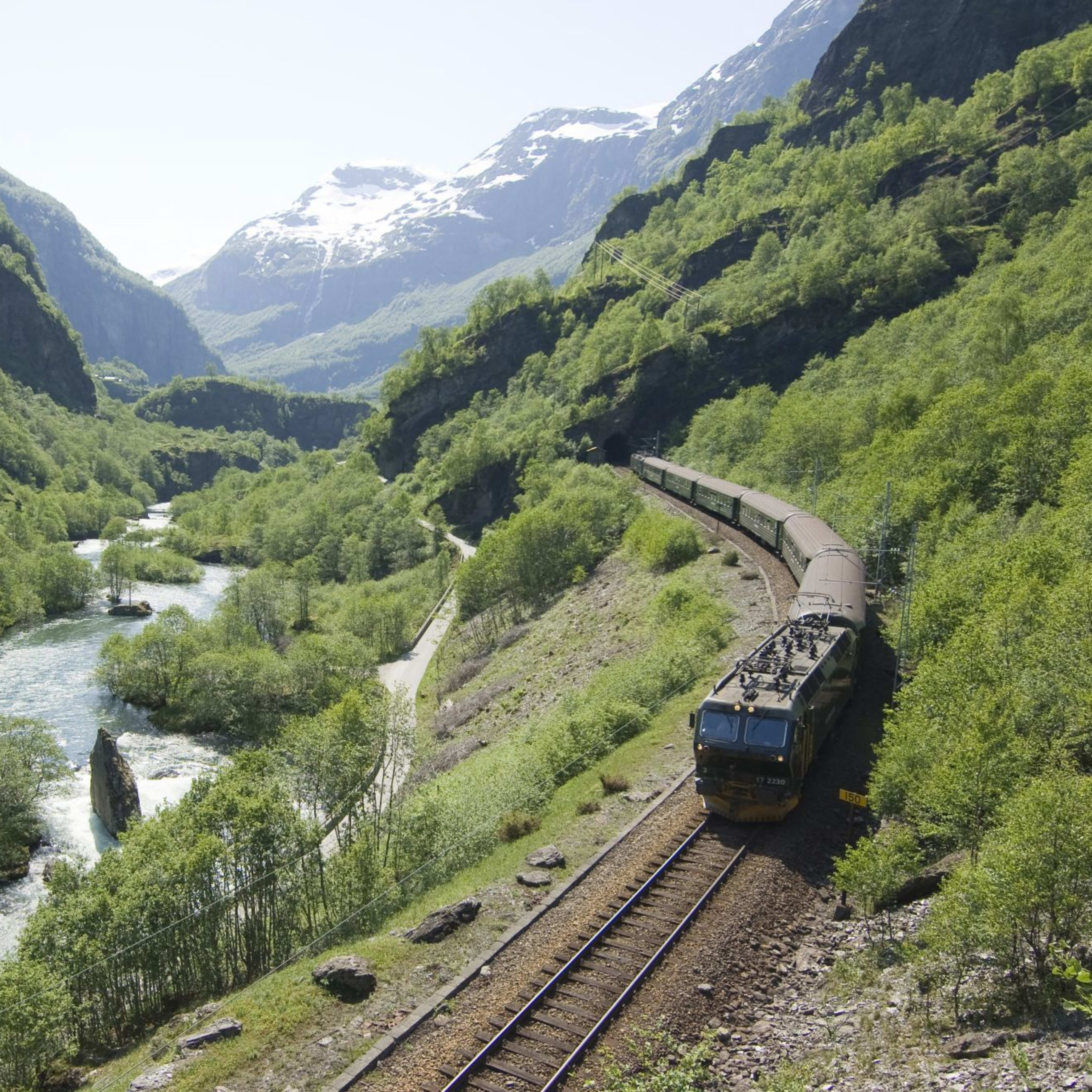 The scenic Flåm railway rides through green valley, river and snowy peaks in view.
