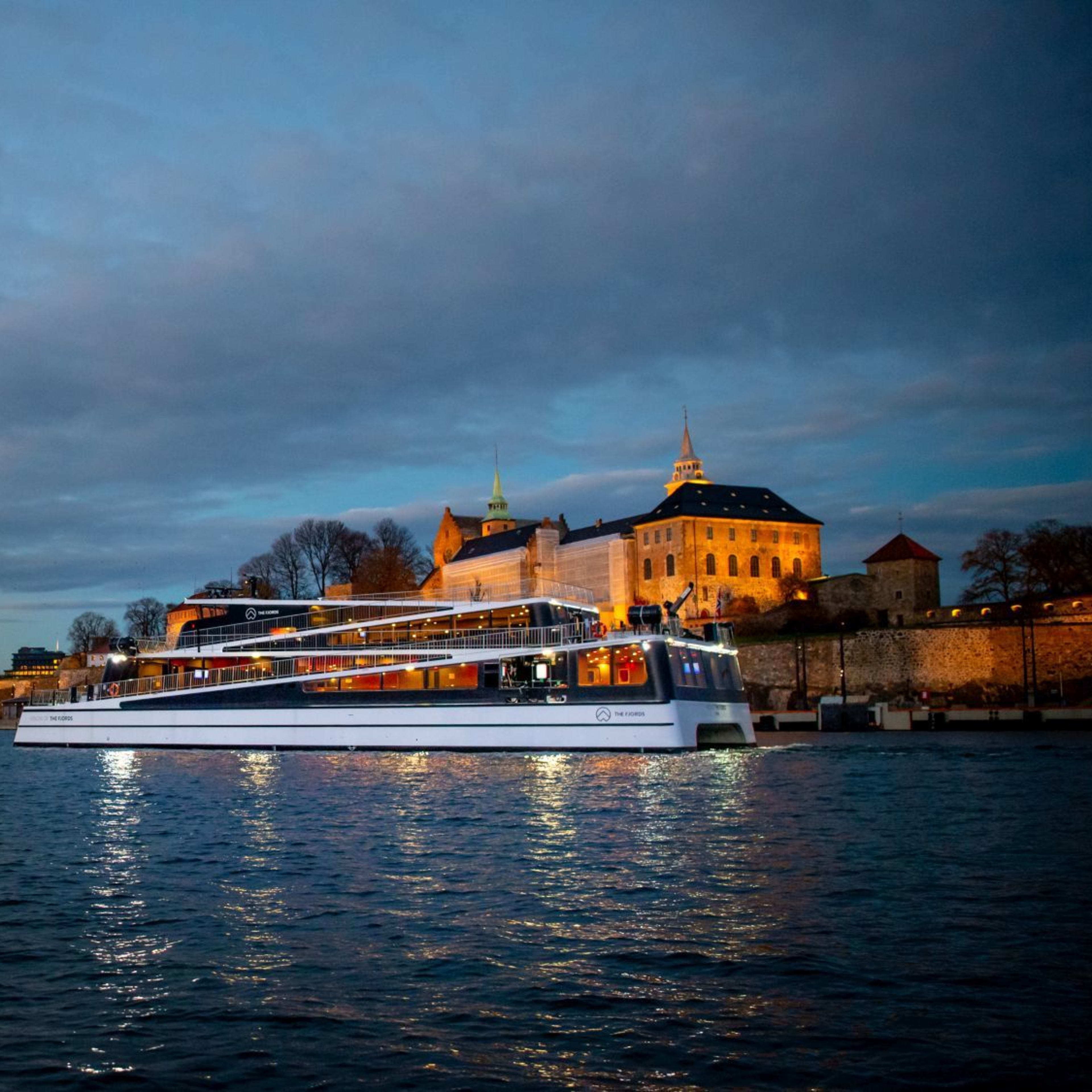 Sleek ferry at dusk near glowing Akershus, warm skyline, and dark clouded sky.