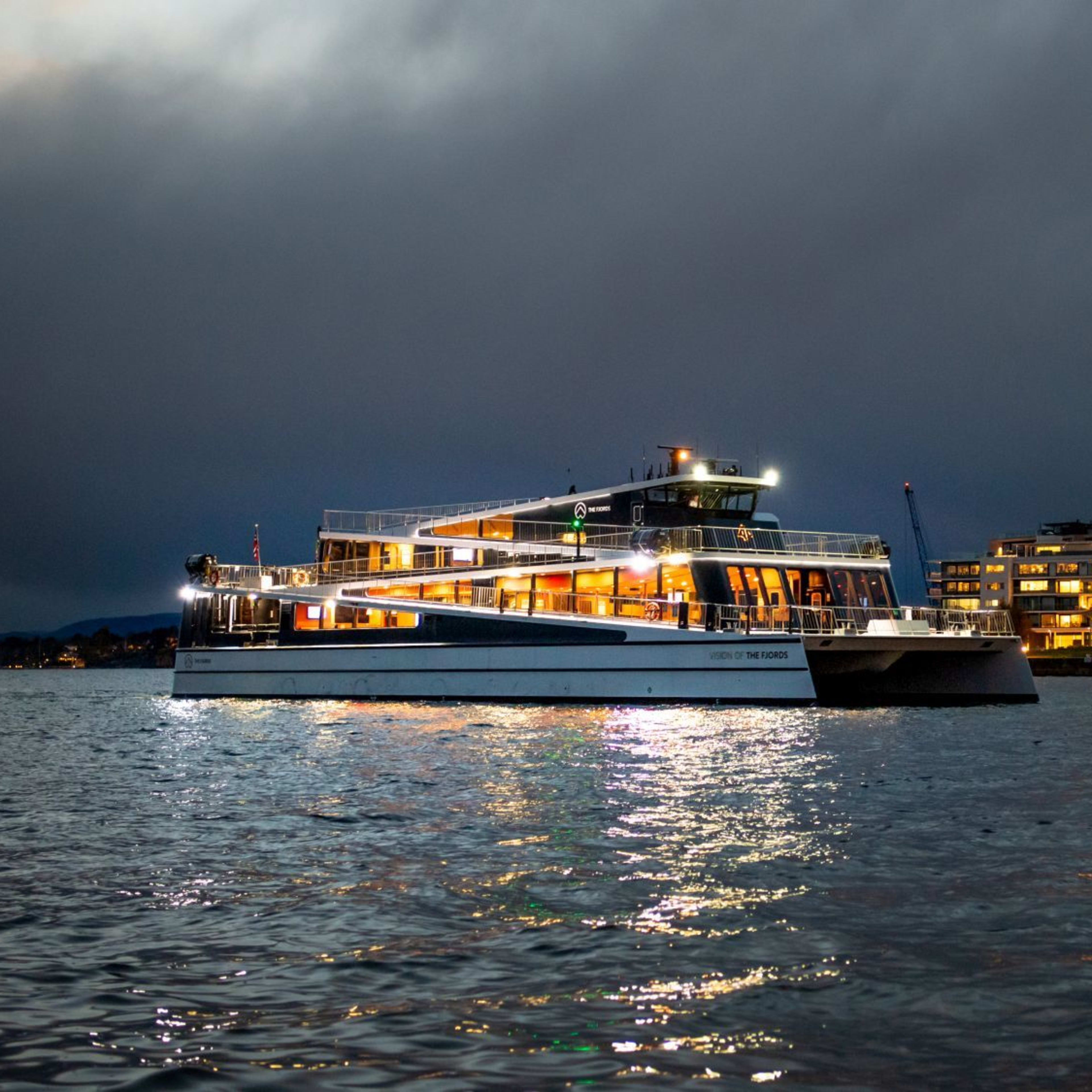 "VISION OF THE FJORDS" ferry sails at night past modern buildings and dramatic sky.