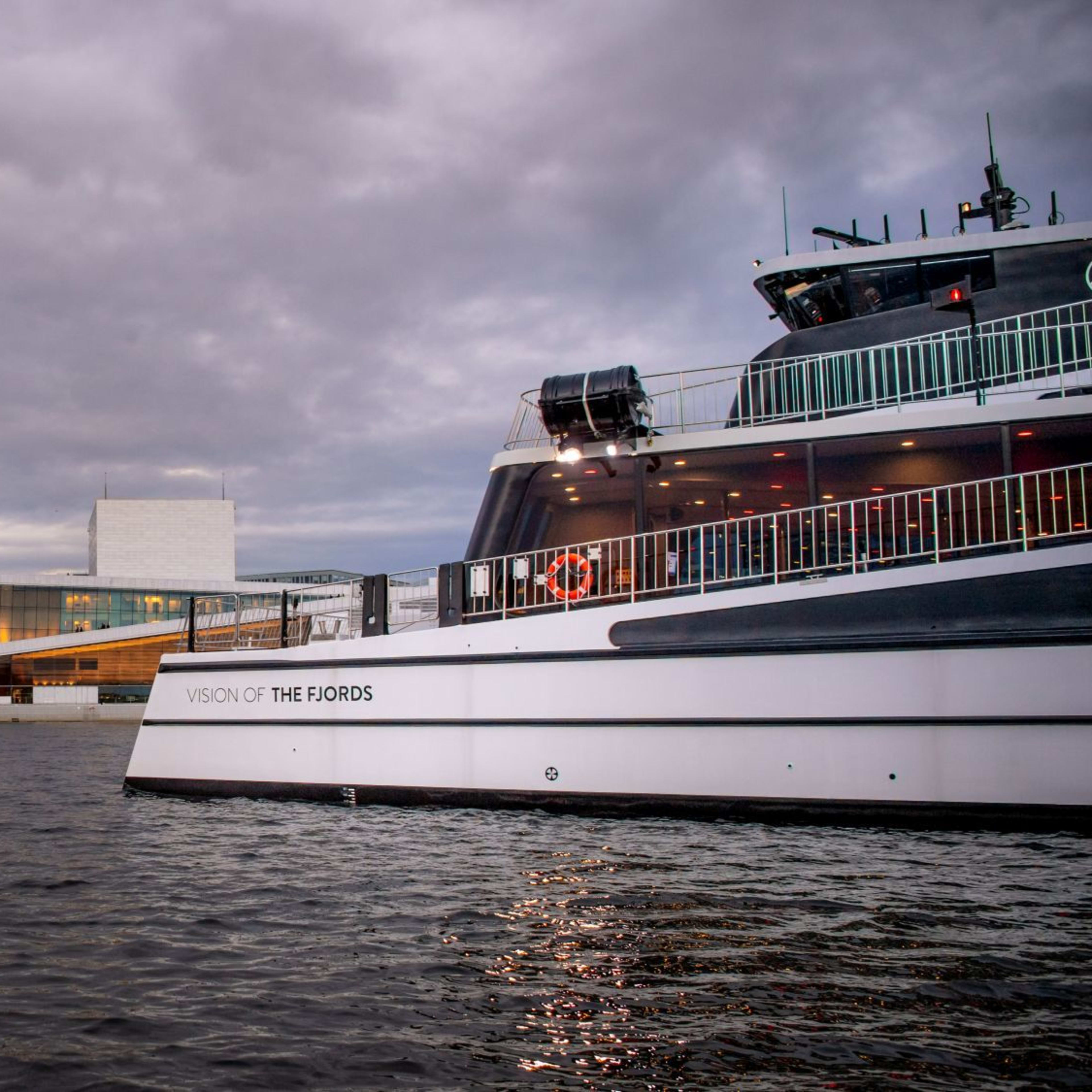 "VISION OF THE FJORDS" ferry passes modern glass building, city skyline under moody sky.