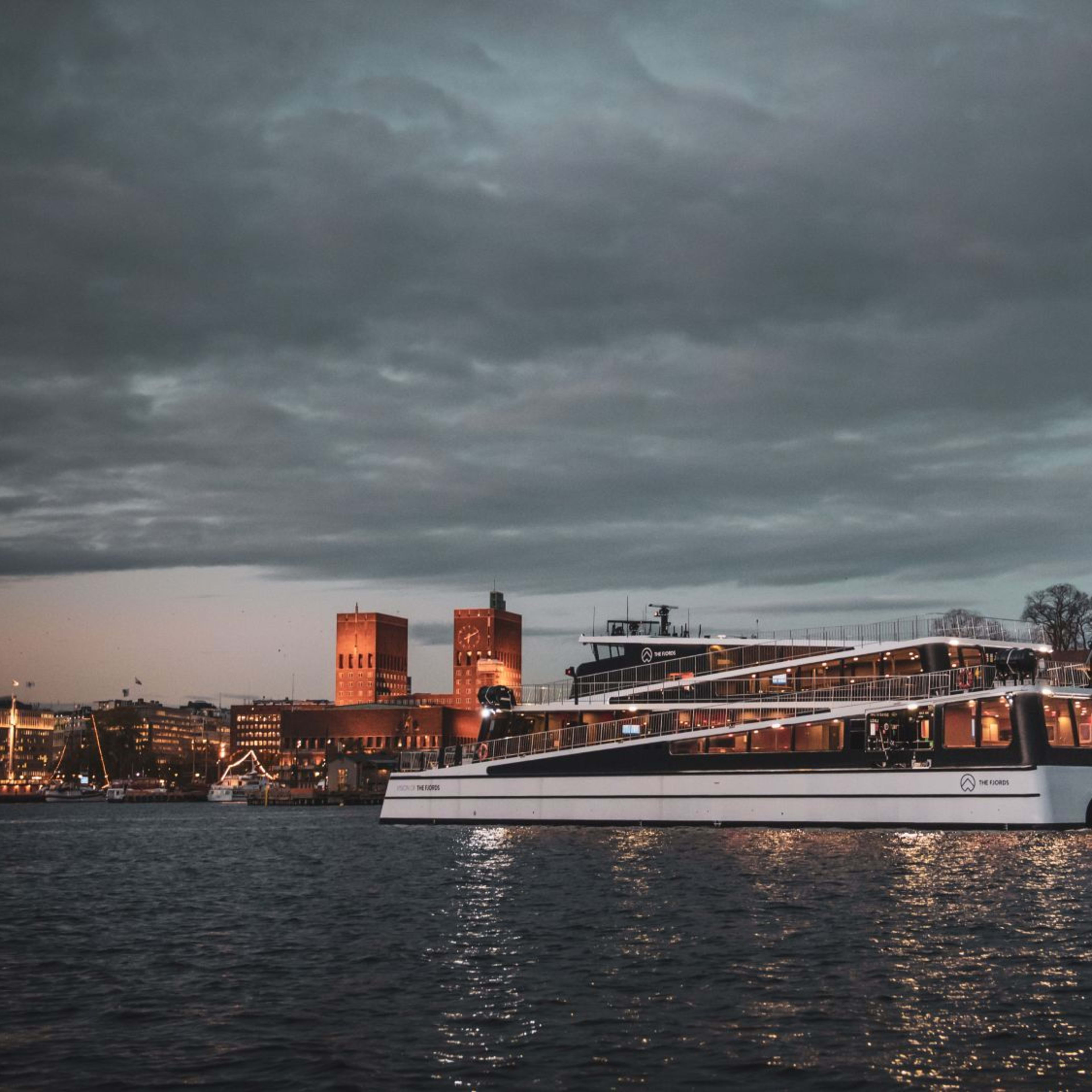 Night ferry passes Akershus Fortress with city skyline aglow and reflections on water.