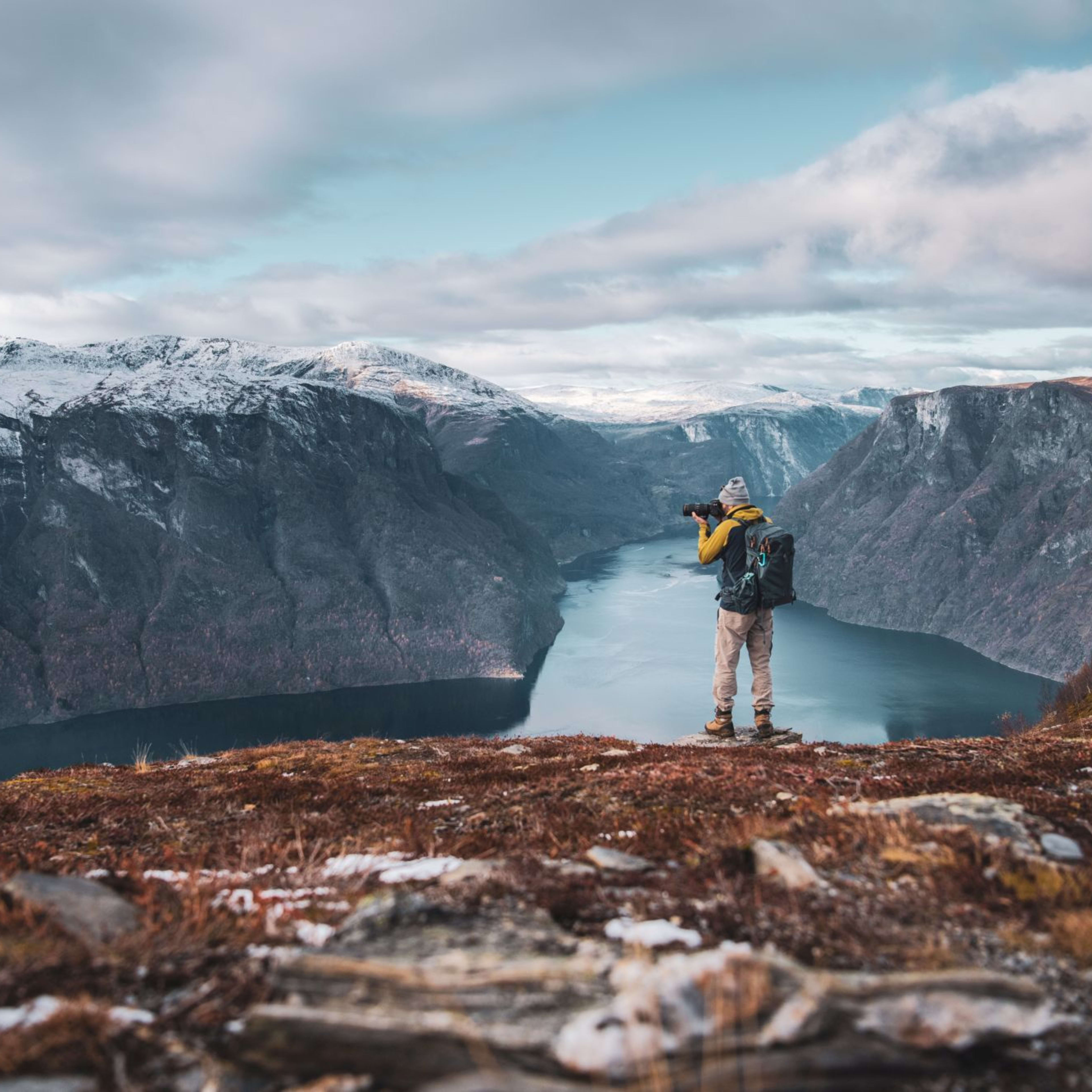 Hiker on rocky cliff with camera, panoramic view of Aurlandsfjord and snow-capped mountains.