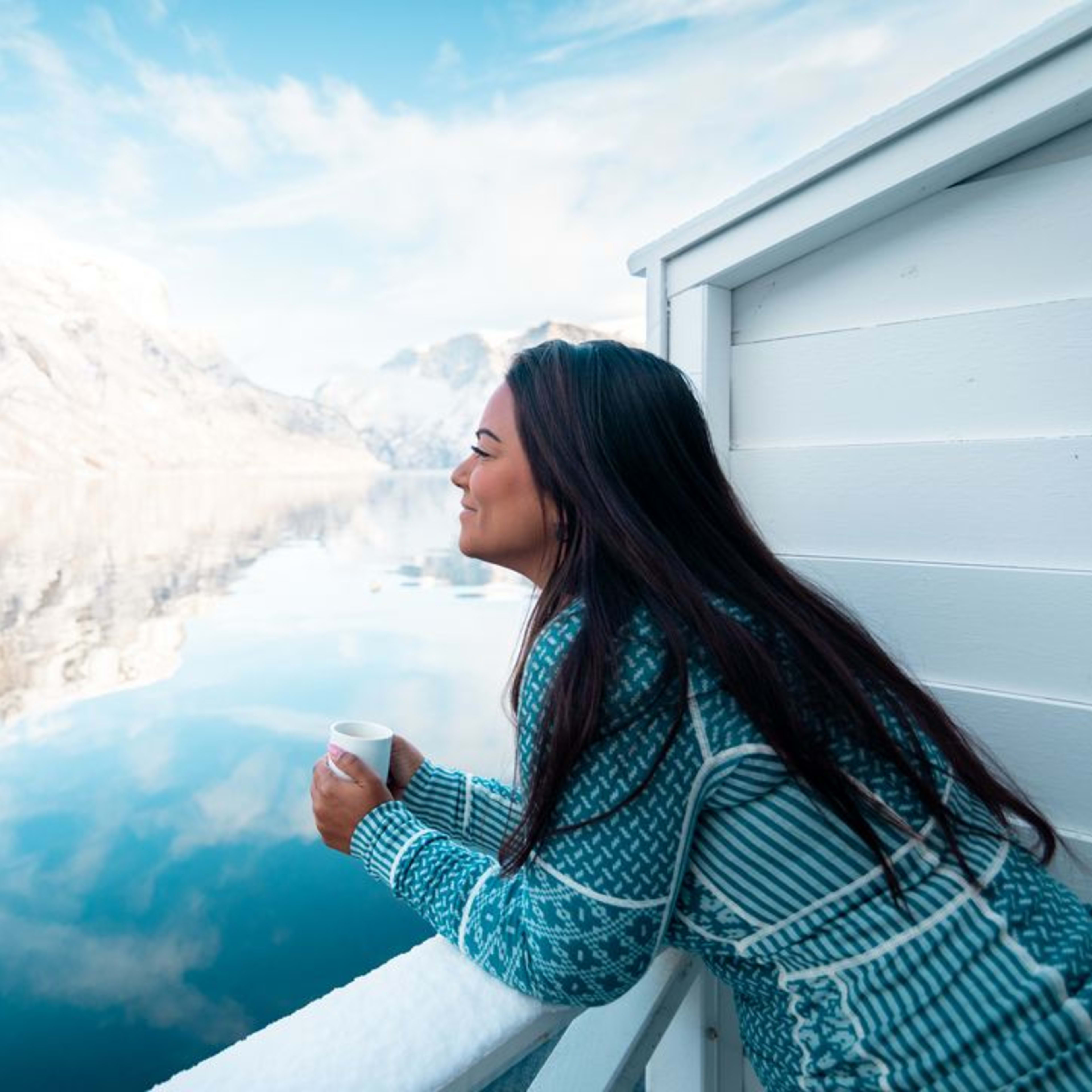 Kvinne i genser holder kopp og ser på snødekt Nærøyfjord og fjell.