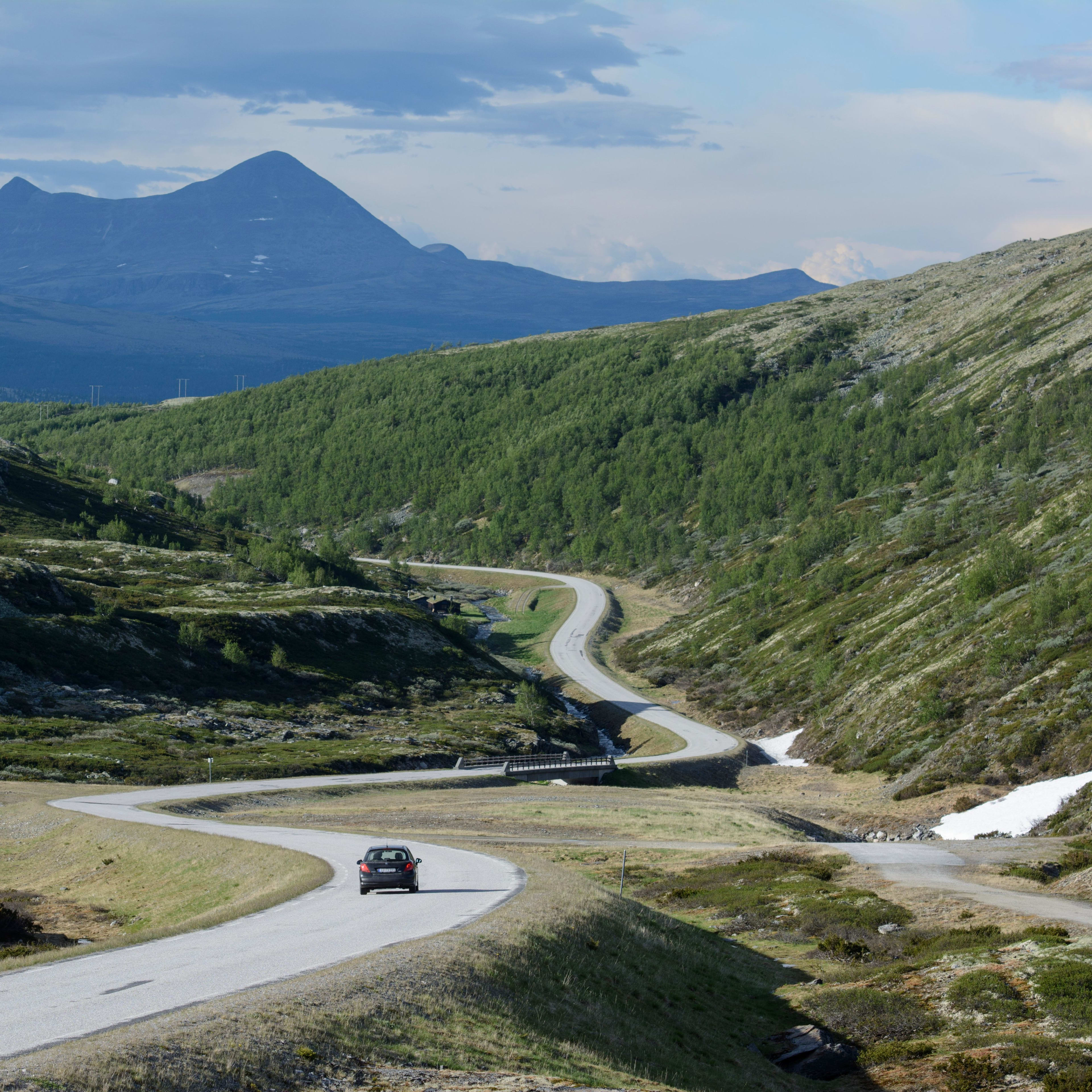 Car on winding road through green valley with rocky hills and distant peaks.
