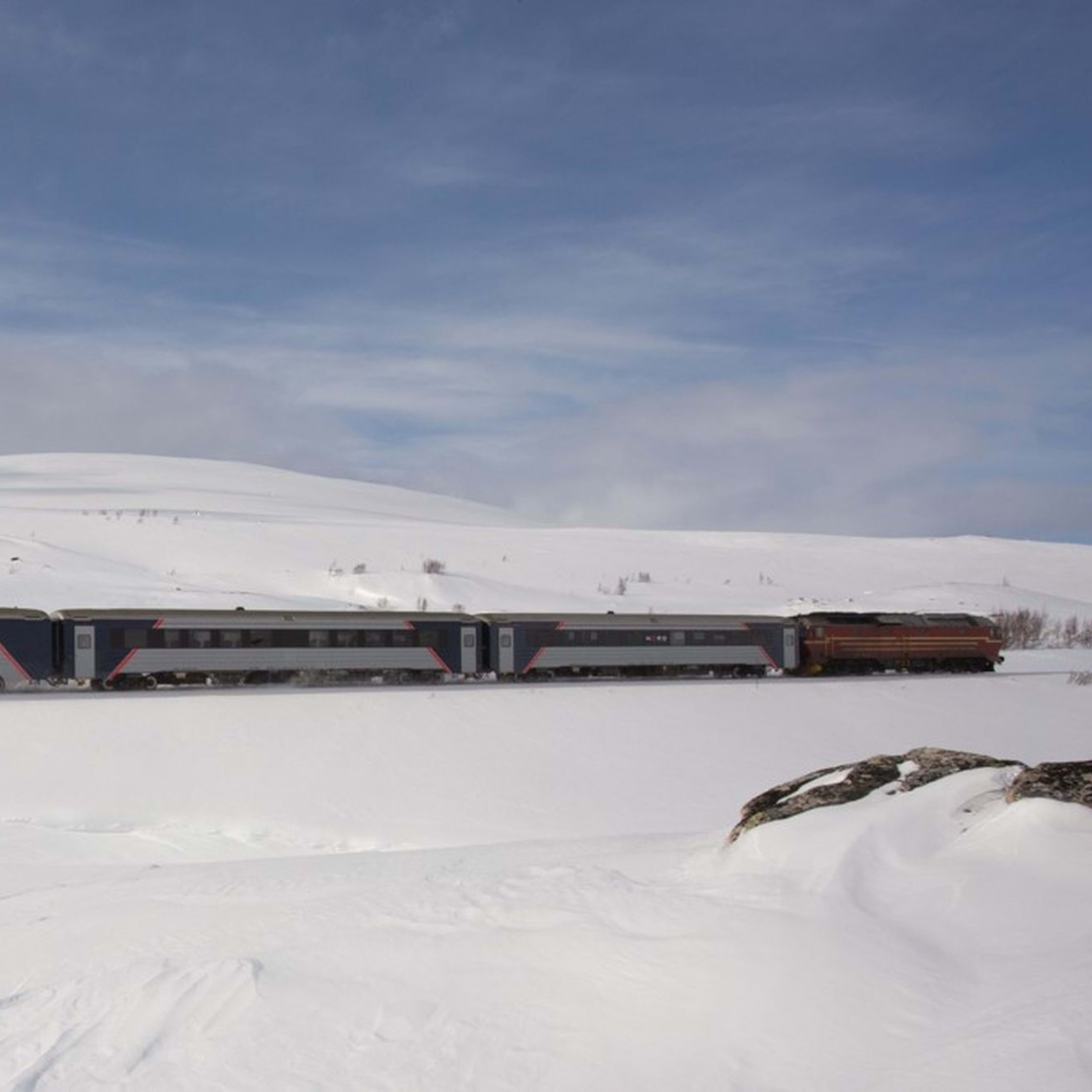 Passenger train through snowy plains and gentle hills under cloudy sky.