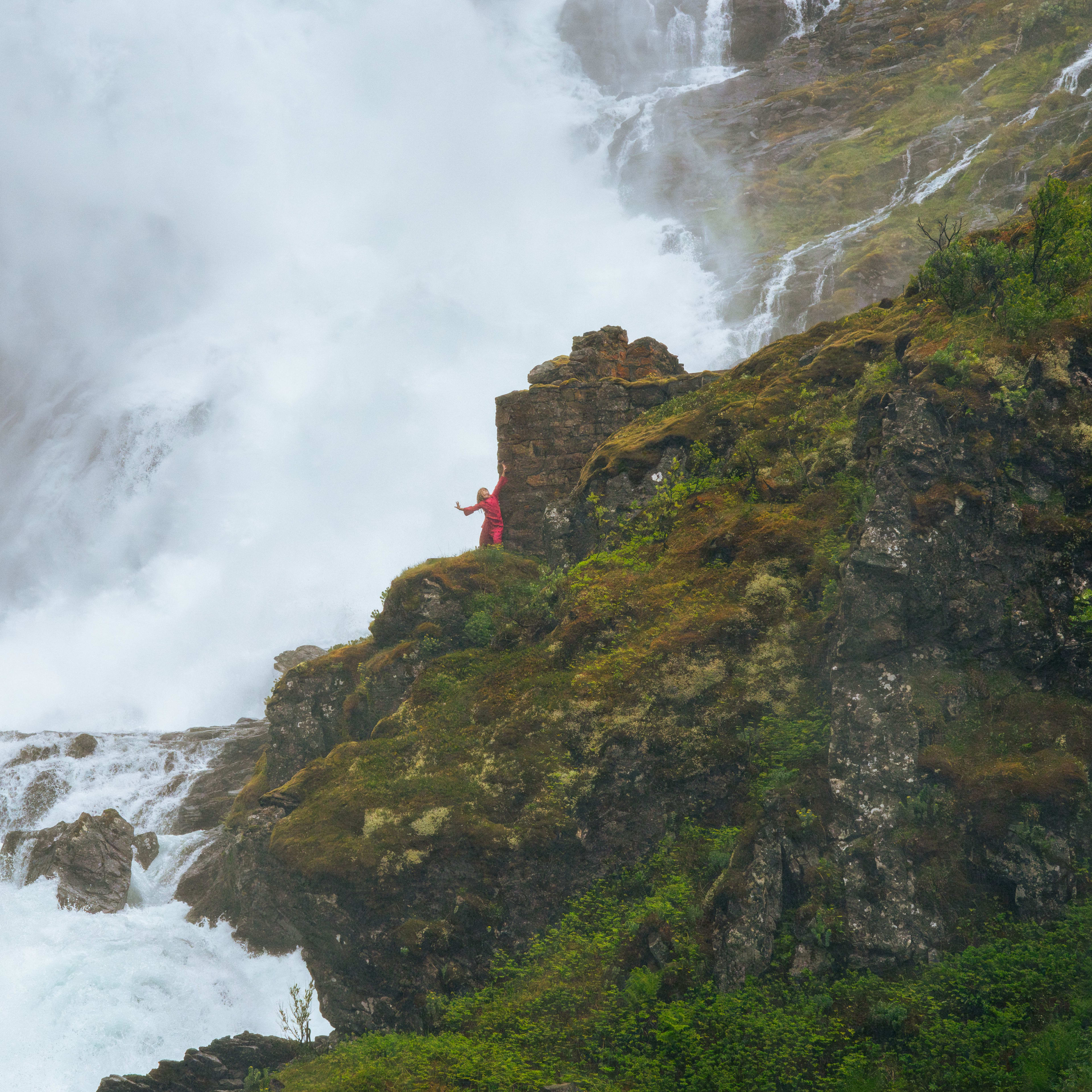 Person i rødt står på mosekledd klippe ved tåkelagt foss i grønn kløft.