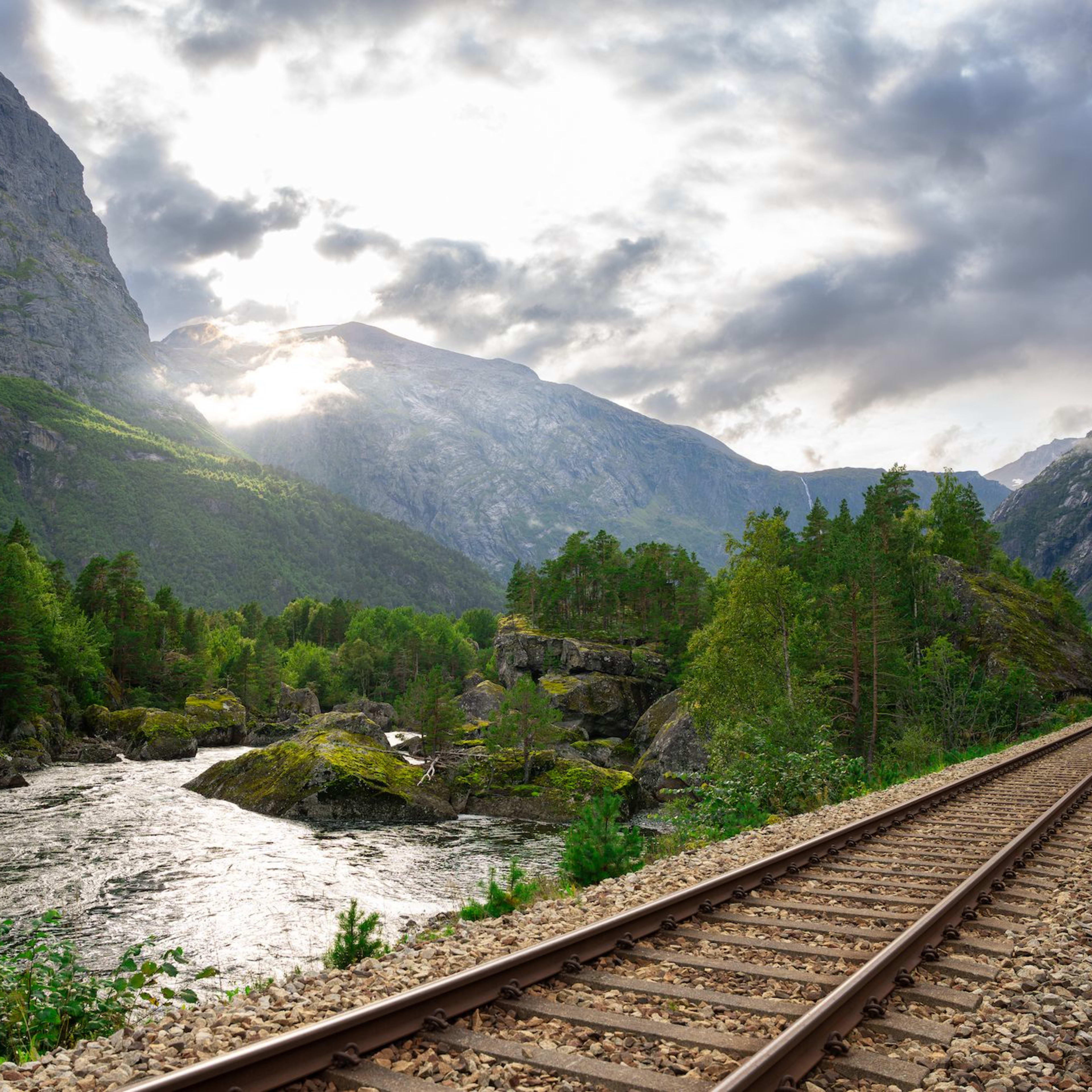 Ein malerischer Blick auf ein Bahngleis, das sich durch ein Tal mit einem Fluss und üppigen grünen Bergen schlängelt, wobei Sonnenlicht durch Wolken filtert.