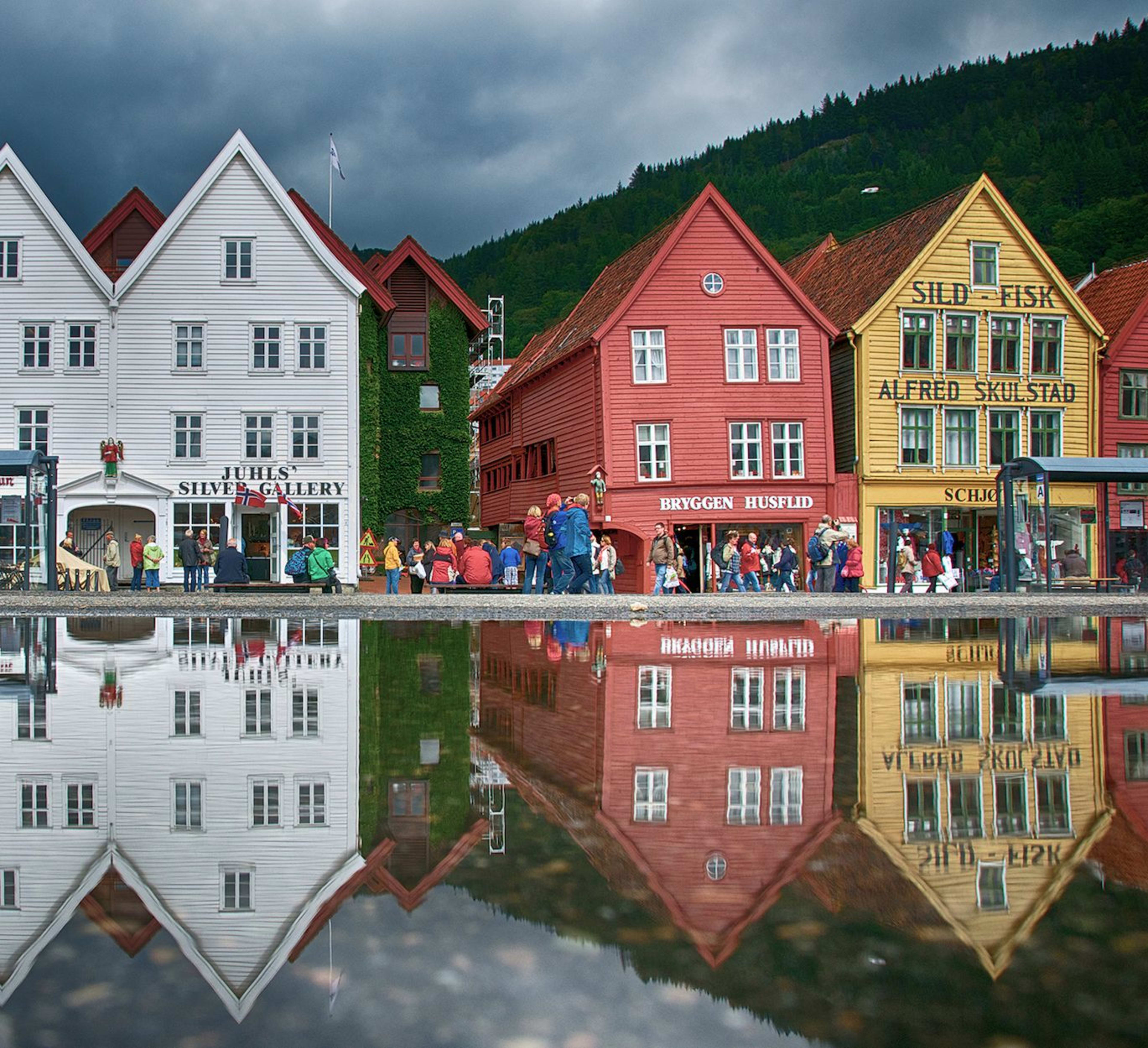 Fargerike Bryggen-bygninger i Bergen speiles i en hav, folk går under dramatiske skyer.