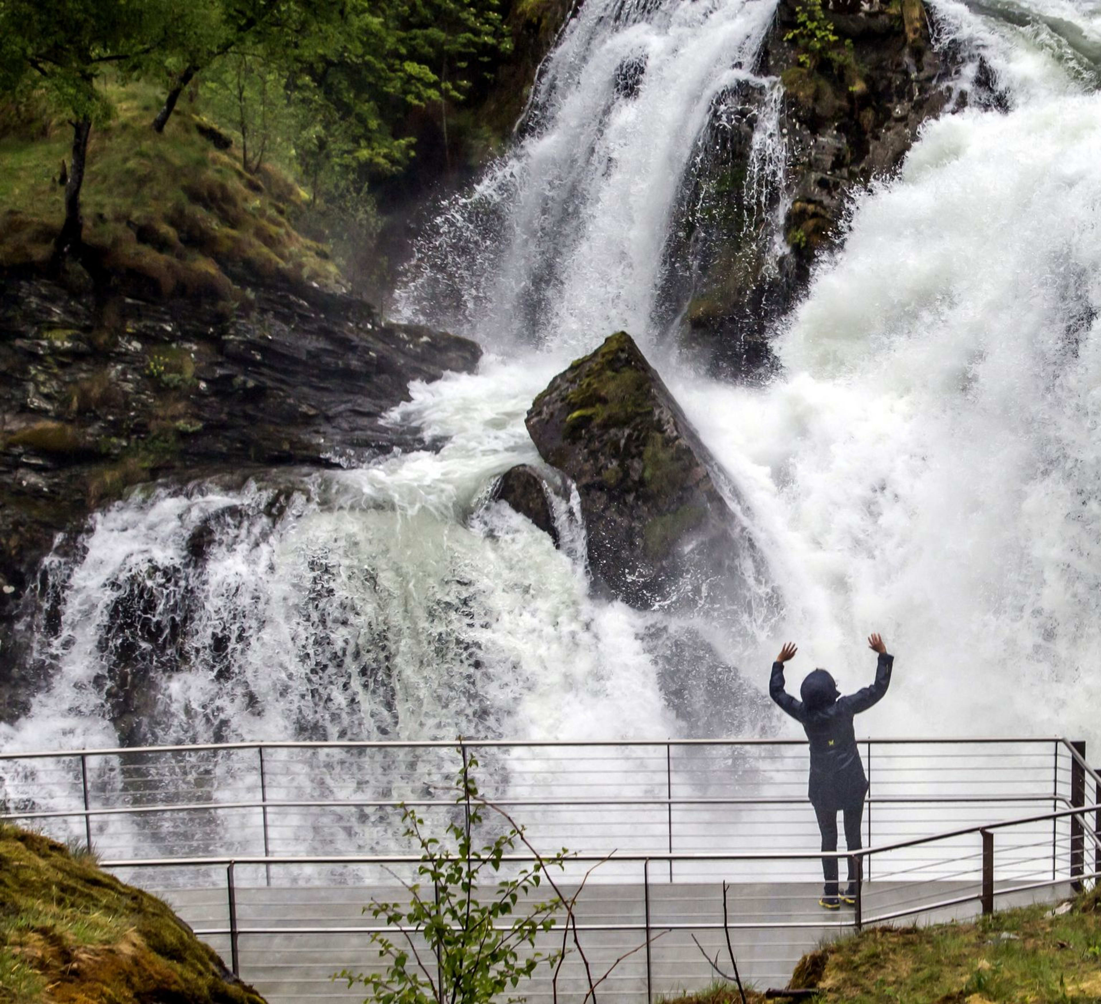 Person in rain jacket raises arms facing massive waterfall among greenery.