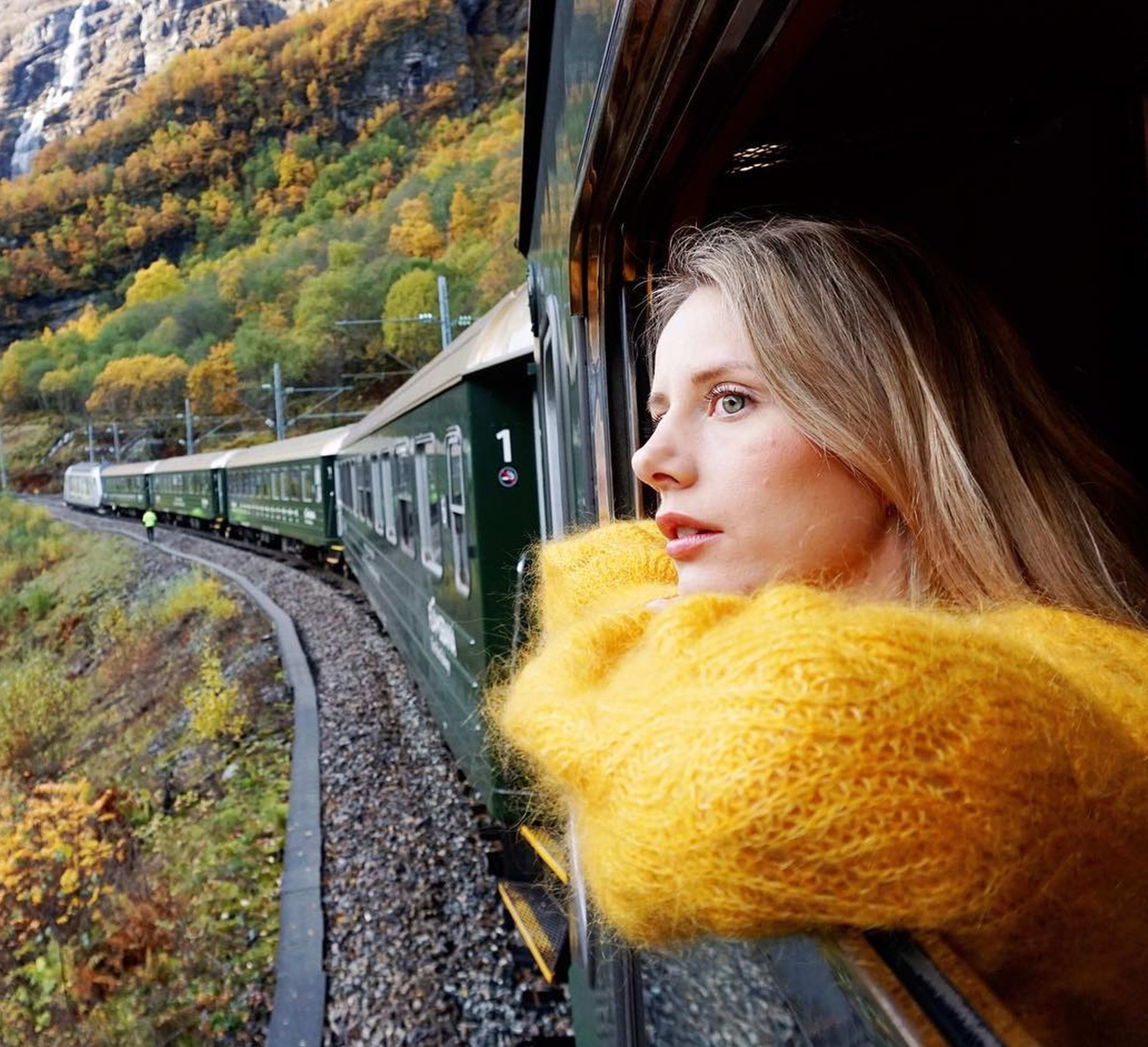 Frau im gelben Pullover blickt aus grünem Zugfenster auf herbstliche Landschaft.