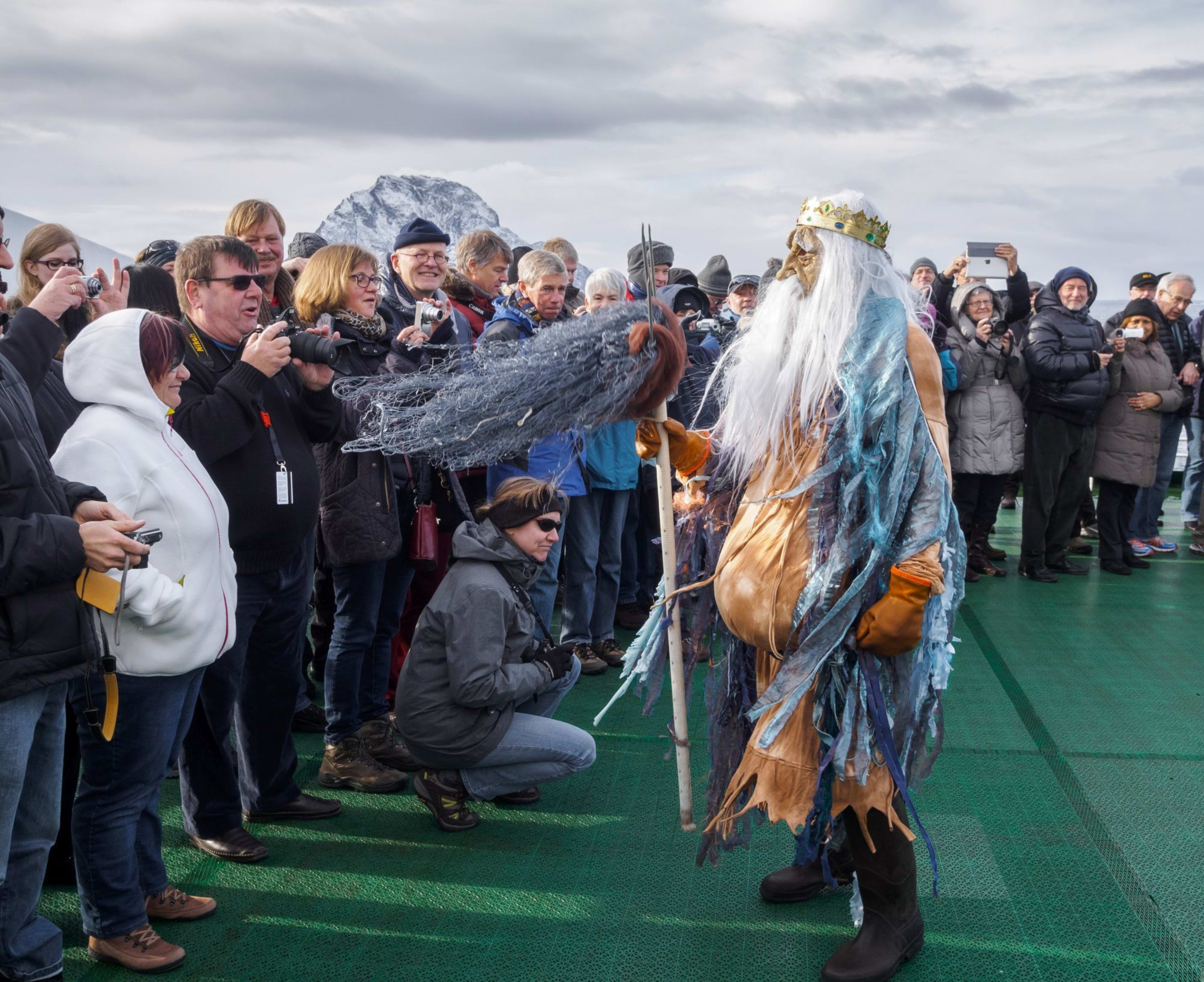 Kostymert performer som sjøkonge med hvitt hår, gullkrone og blå kjole, underholder publikum på ferjedekk med snødekte fjell bak.