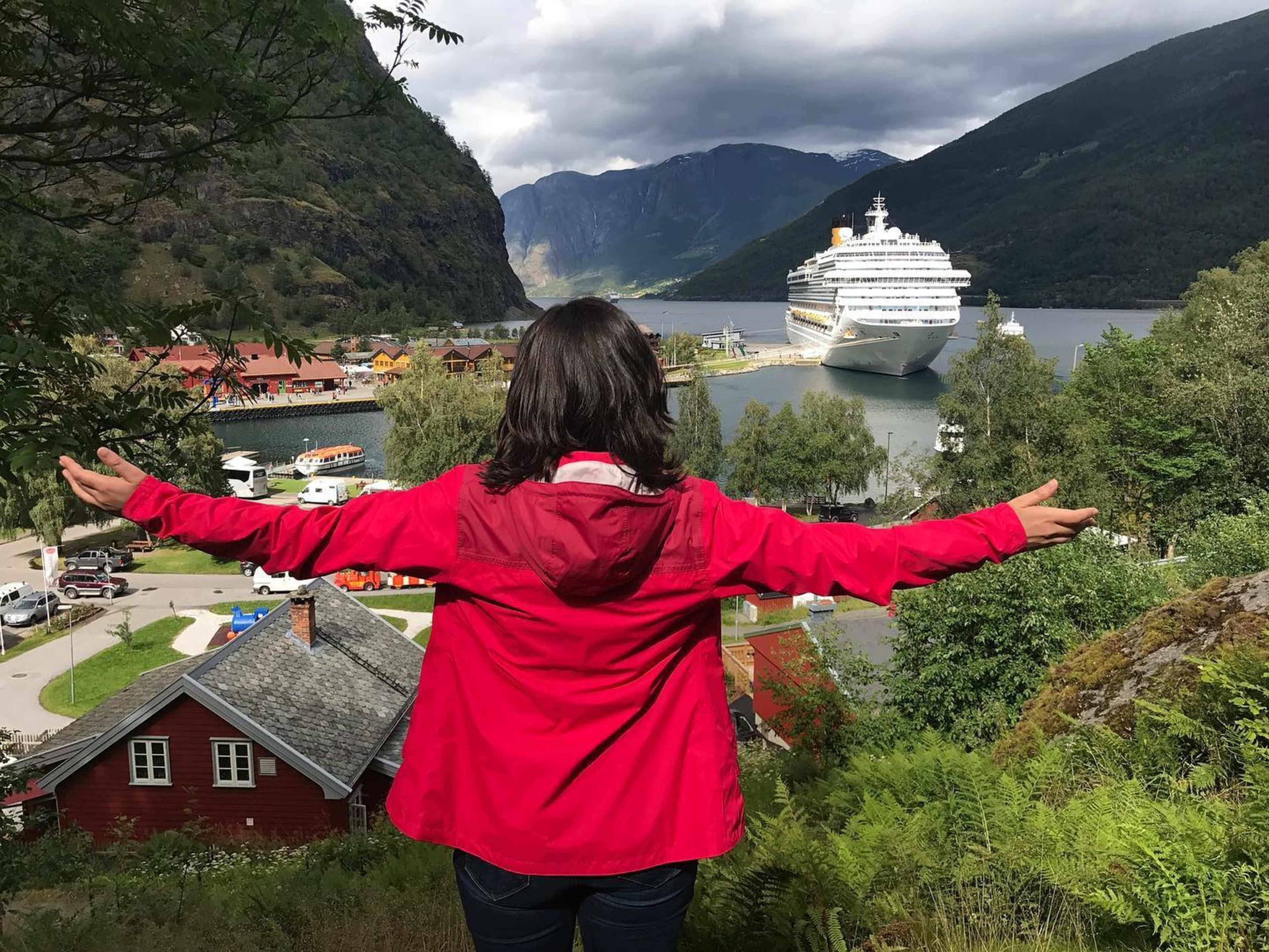 Arms outstretched, facing Flåm and fjord landscape with cruise ship docked at serene port surrounded by hills.