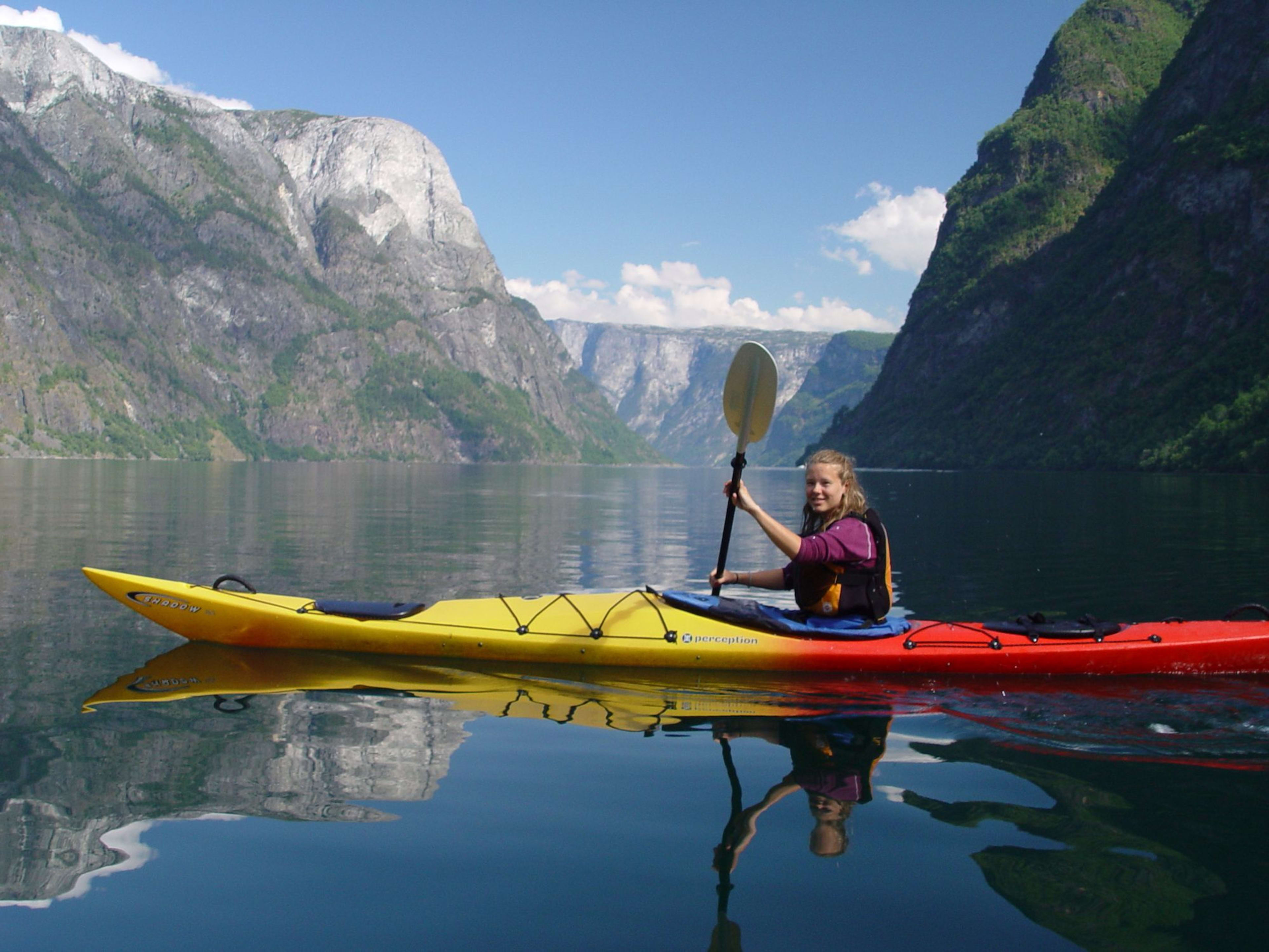 Smiling kayaker paddling a yellow and red kayak on calm water, with cliffs and lush hills surrounding a narrow fjord.