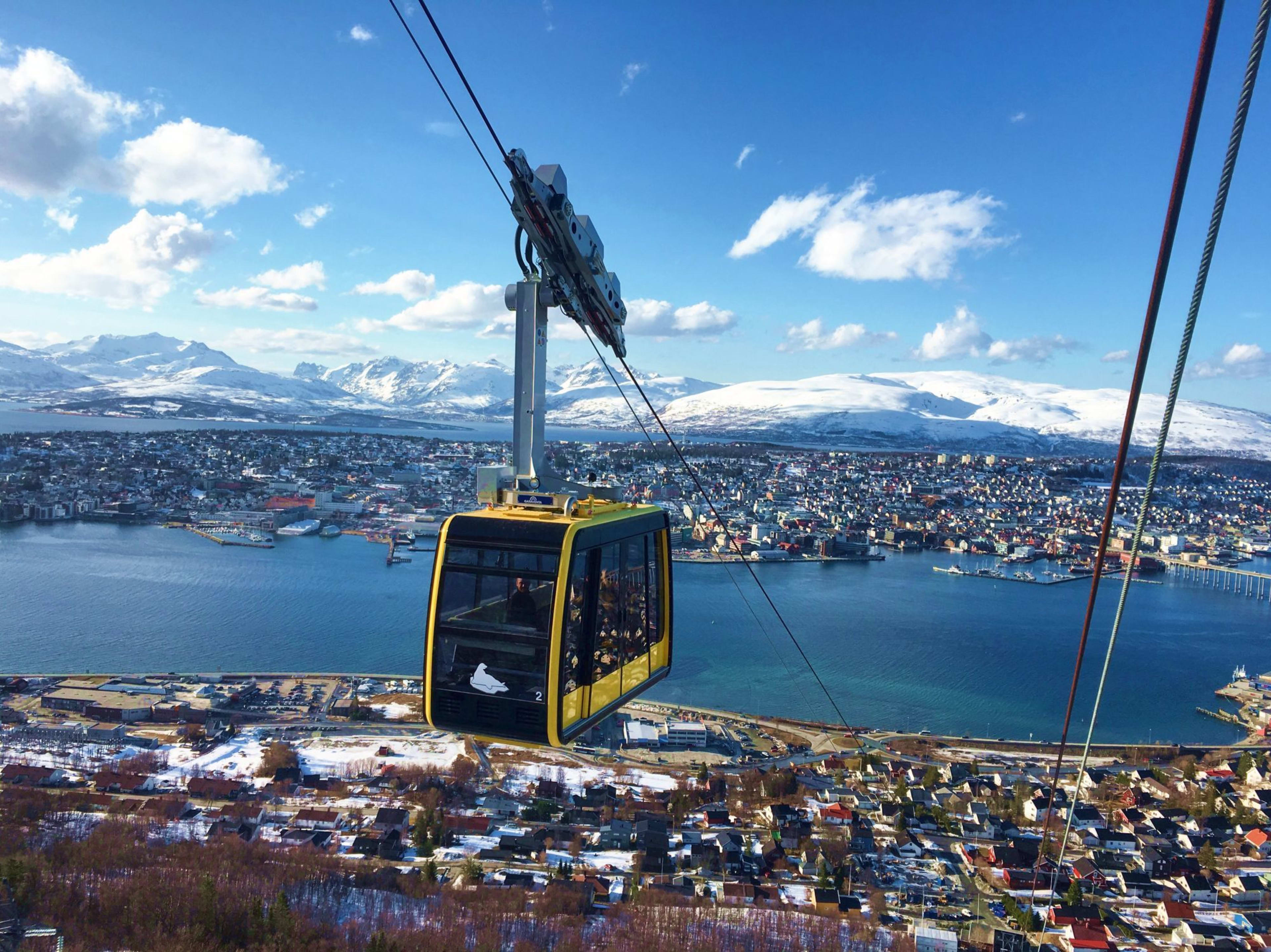 Seilbahnblick über Tromsø mit gelbem Kabin, Hafen, bunten Gebäuden und schneebedeckten Bergen im Hintergrund.