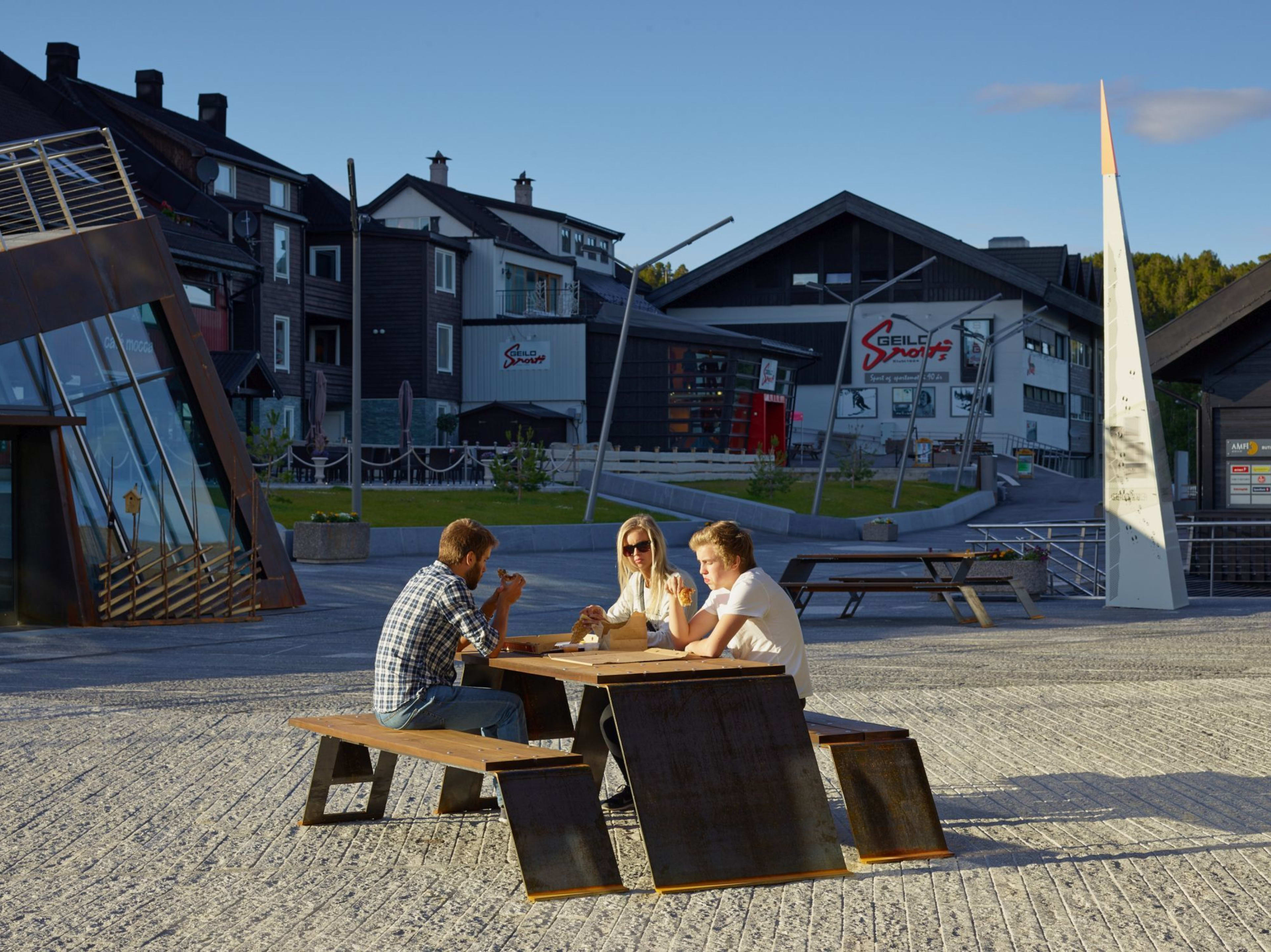 Three enjoy ice cream at picnic table in sunny plaza near alpine shops.