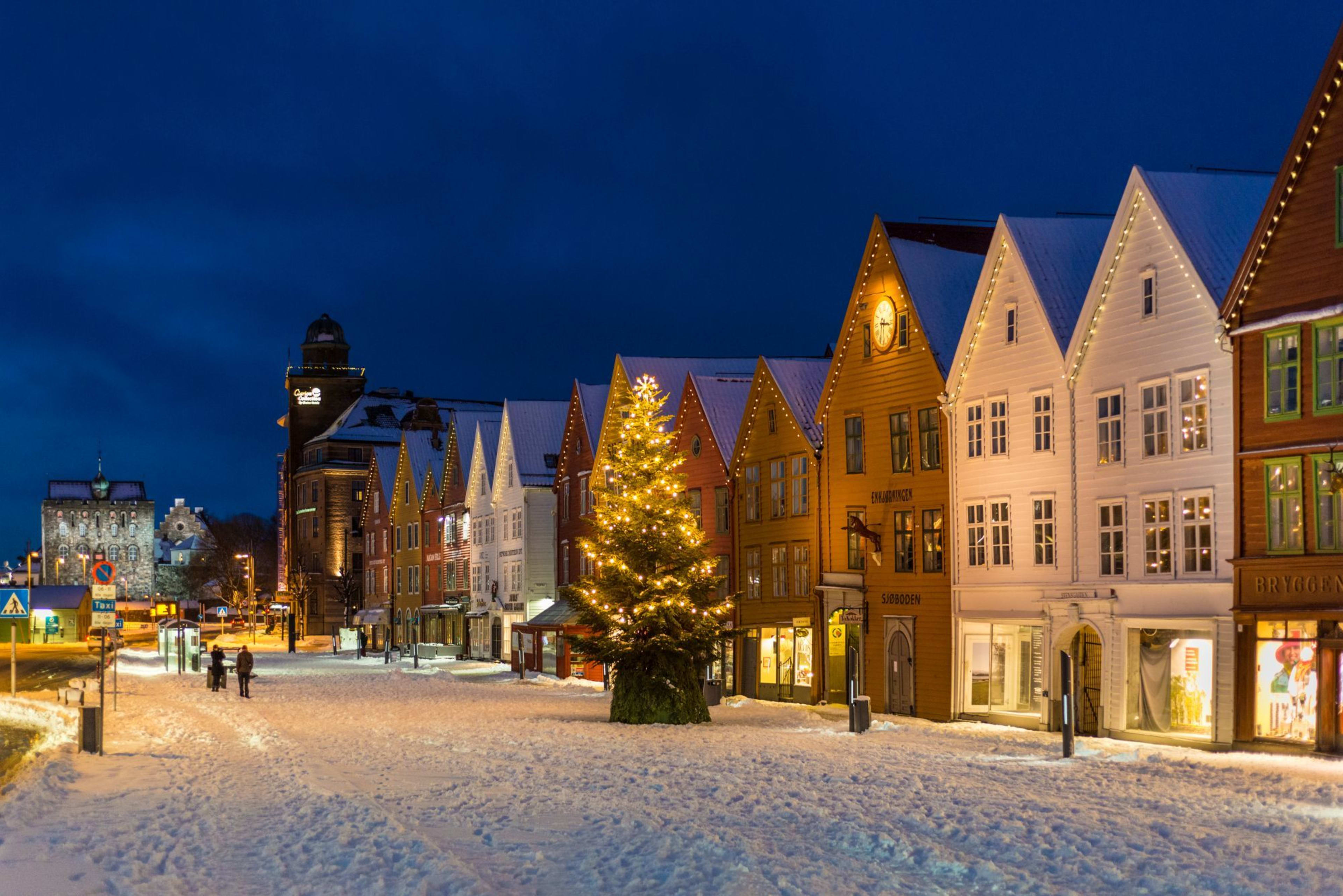 Snow-covered street in Bergen with colorful wooden houses, Christmas lights, and decorated tree creating festive vibe.