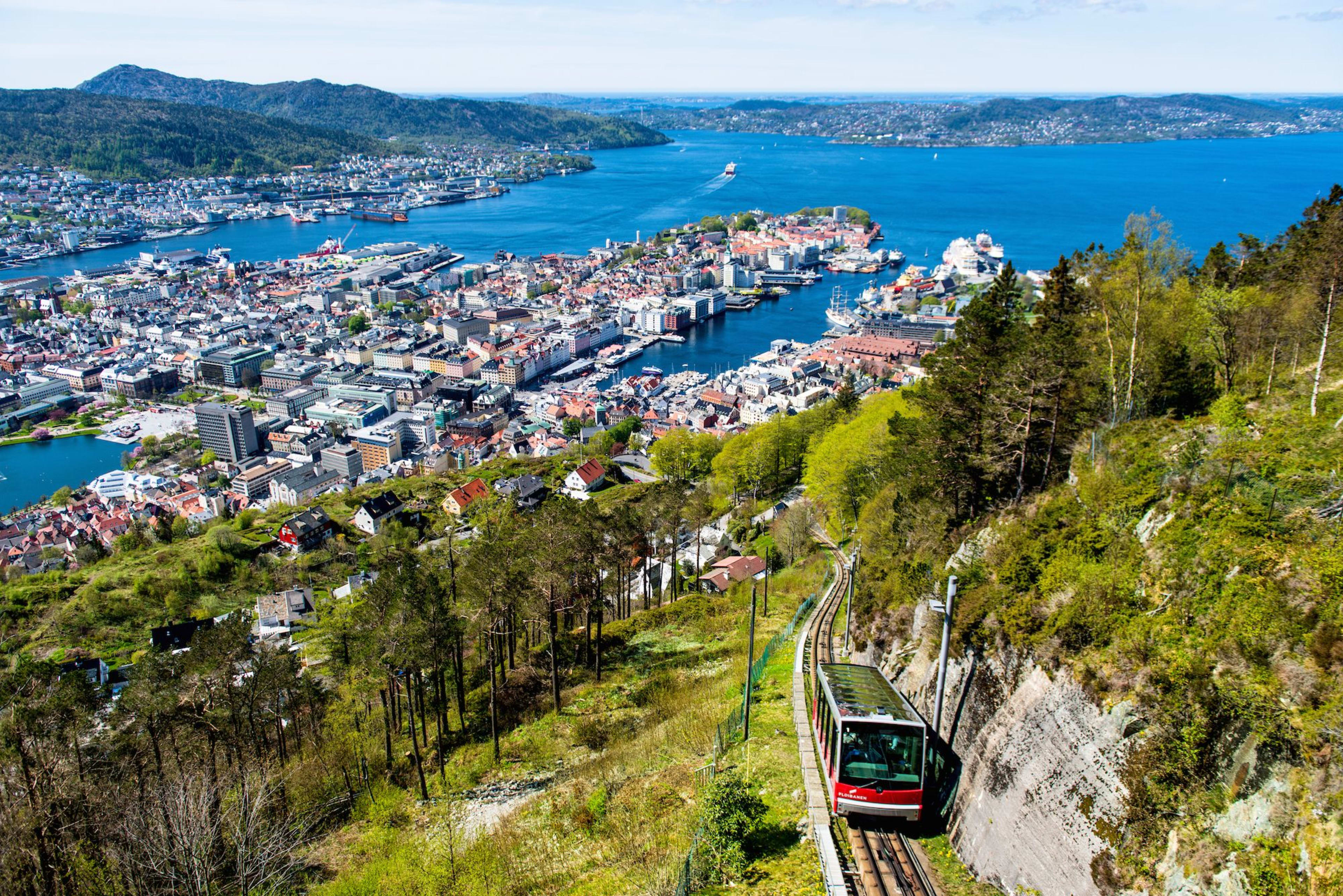 Blick vom Hügel in Bergen mit einer Standseilbahn, die durch Bäume fährt, Stadt, Hafen und farbige Gebäude unter klarem Himmel.