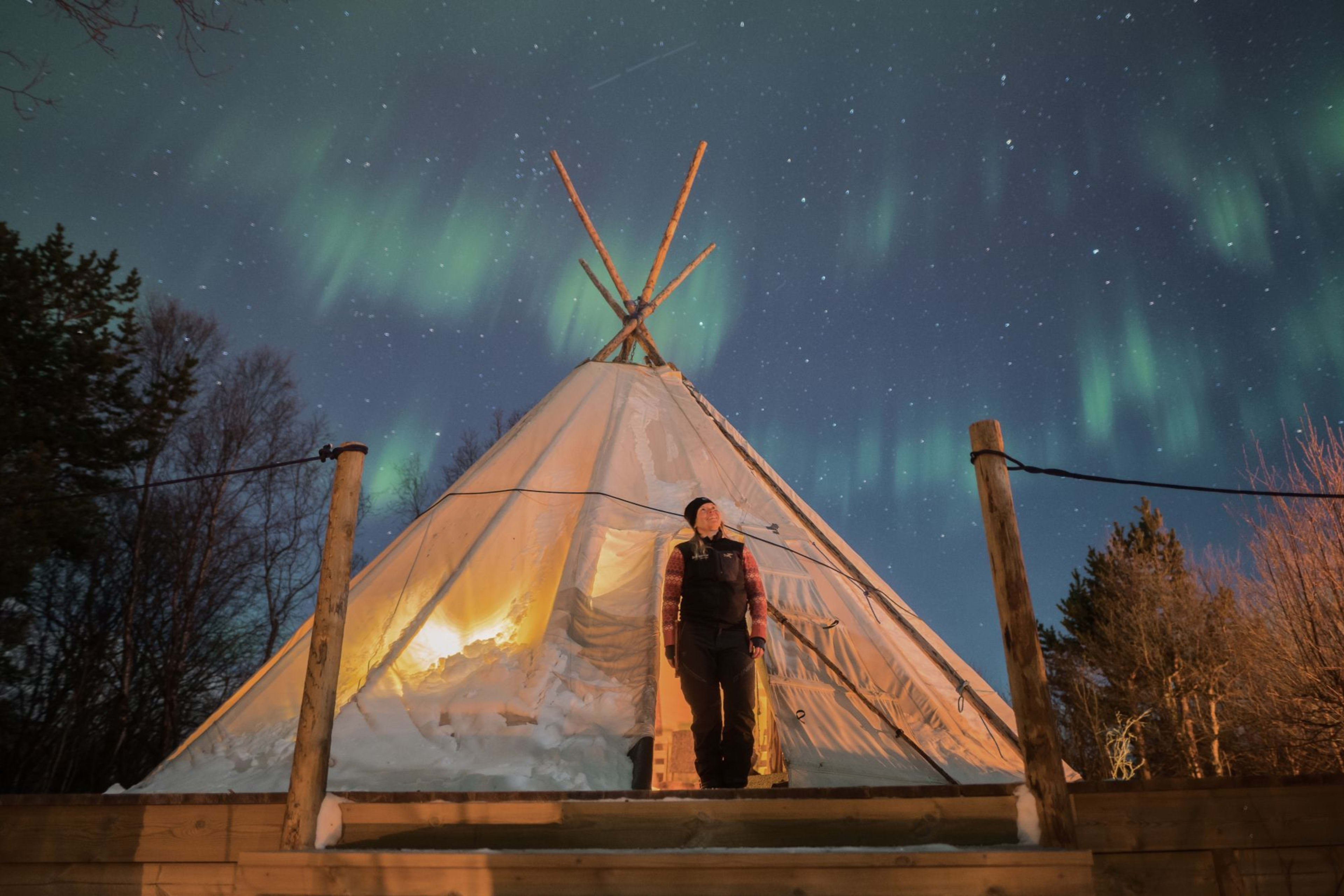 Person standing at a Sami lavvu entrance, gazing up at green Northern Lights in a snowy, serene setting.