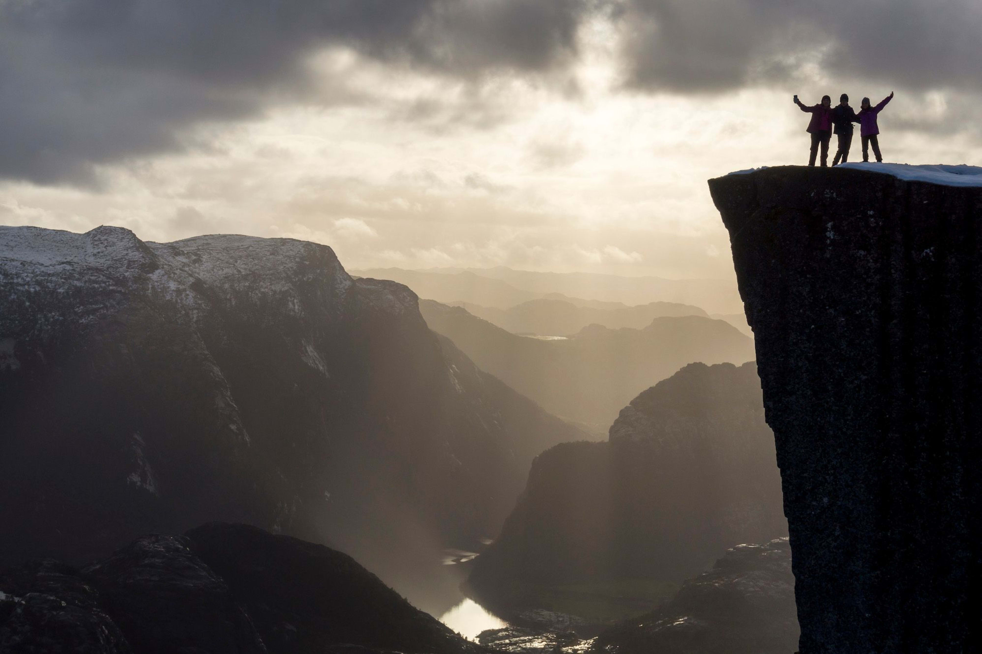 Three silhouetted figures on a cliff edge with golden sunlight breaking through dramatic clouds.