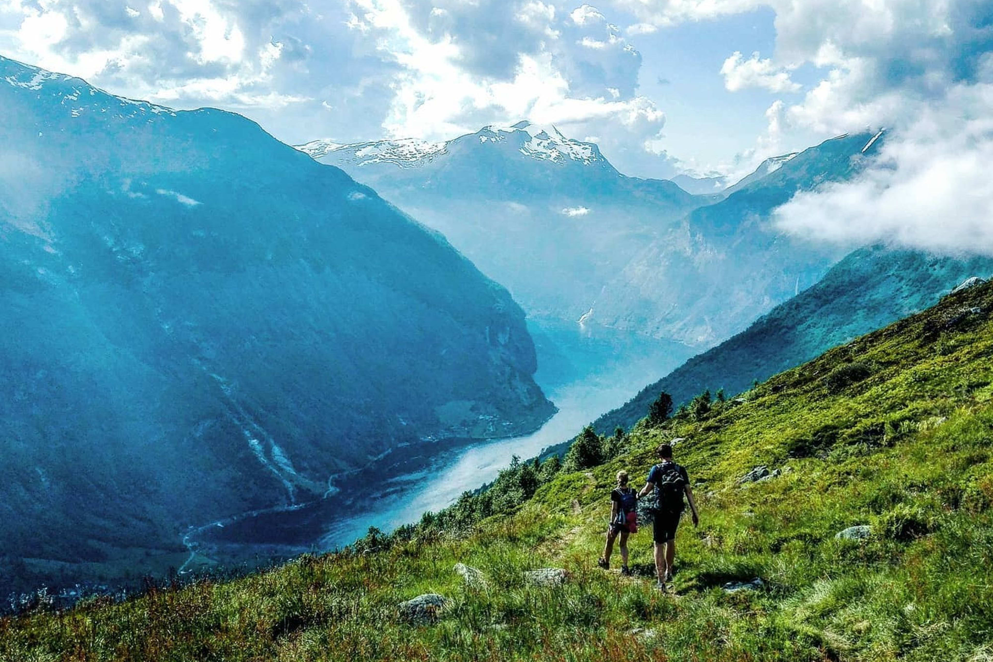 Hikers on trail above misty fjord and tall peaks.