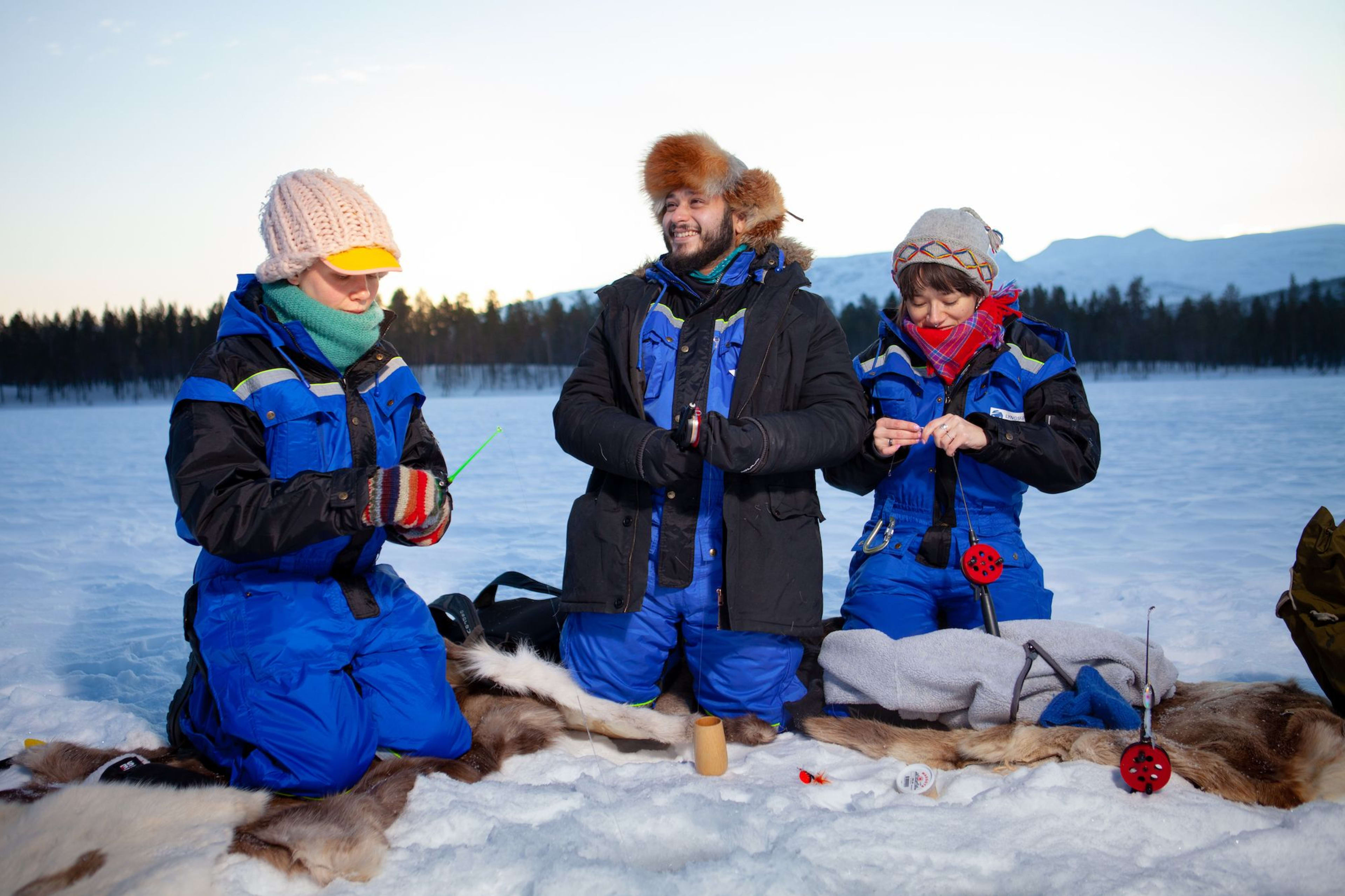 Drei Personen in bunter Winterkleidung bereiten sich auf Eisfischen auf einem zugefrorenen See vor, sitzen auf Rentierfellen, umgeben von Schnee.