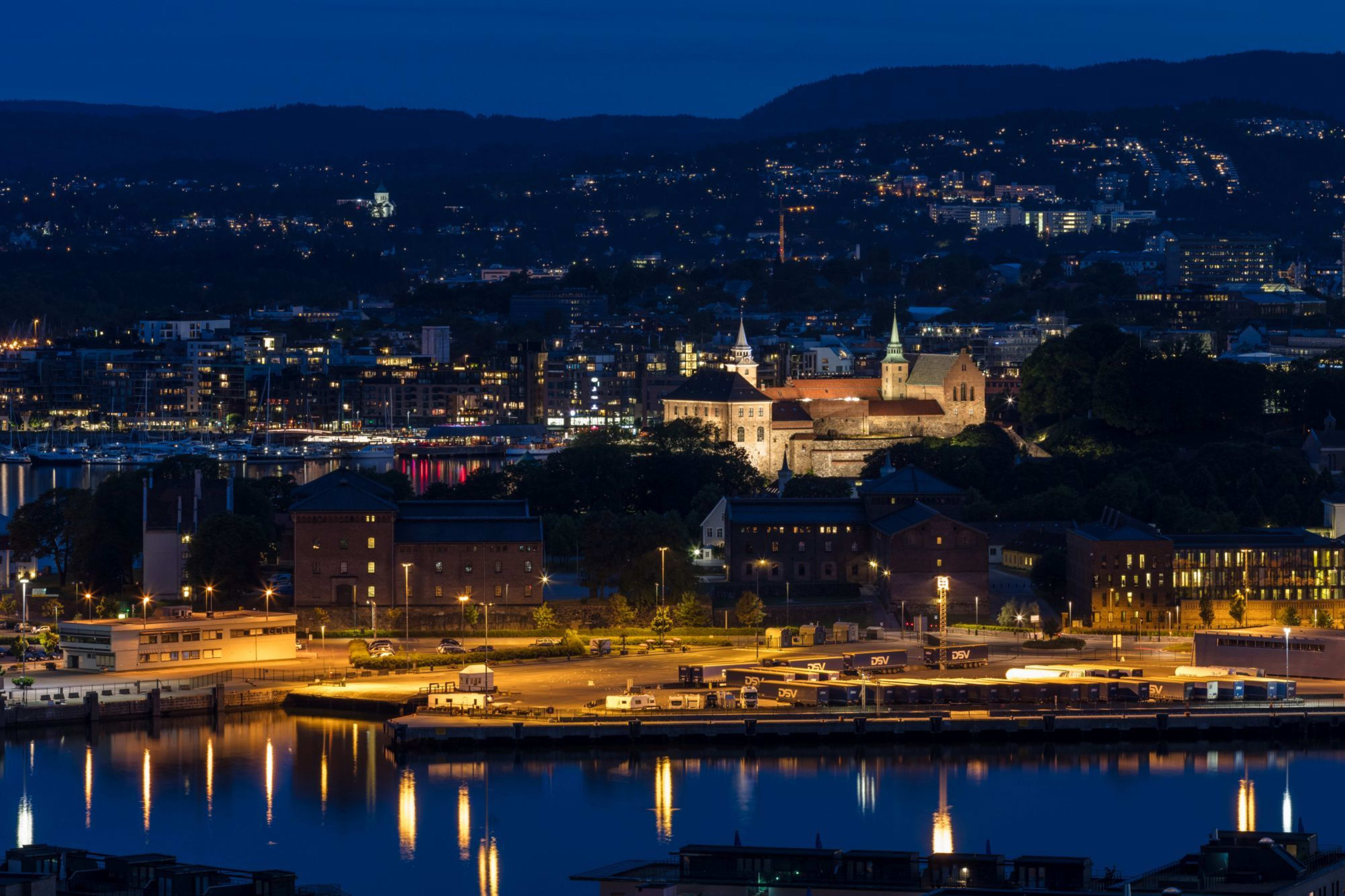 Nattbybilde med historisk slott opplyst, vannrefleksjoner, moderne bygninger og fjerne åser under dyp blå himmel.