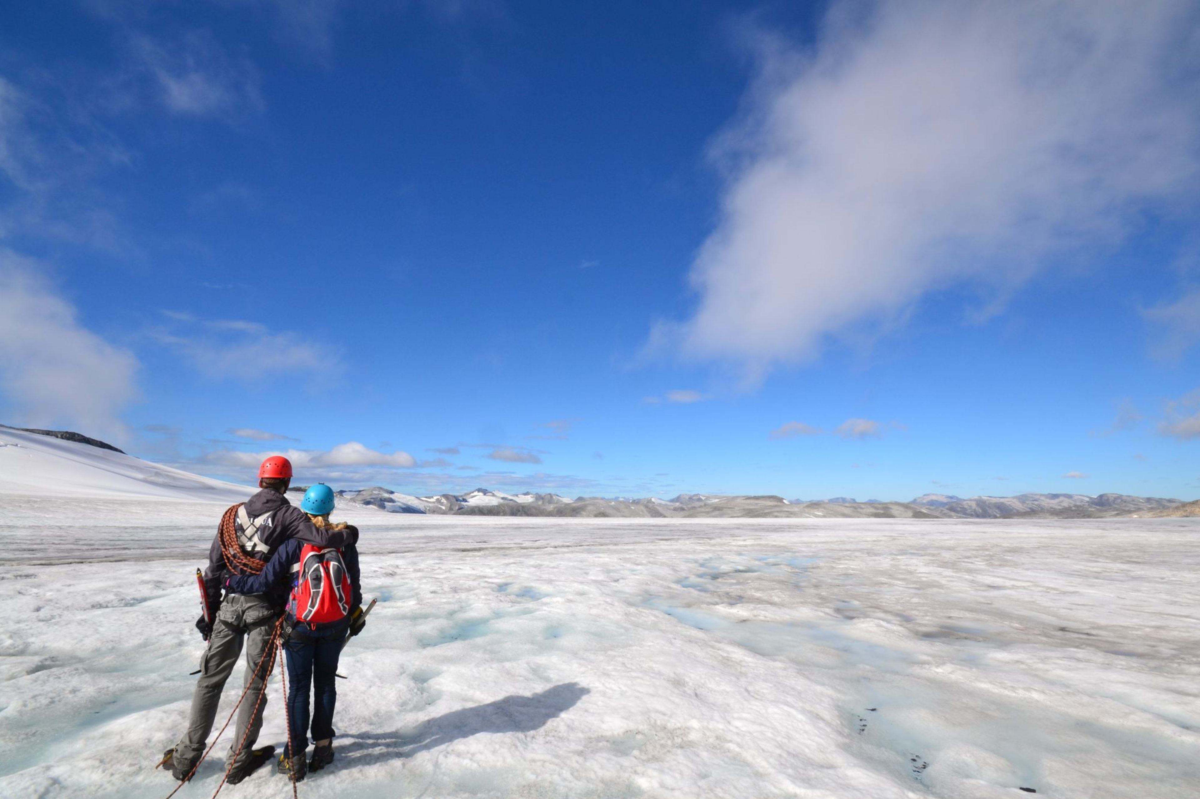Two climbers standing on glacier, one with arm around the other, gazing at snow-covered landscape.