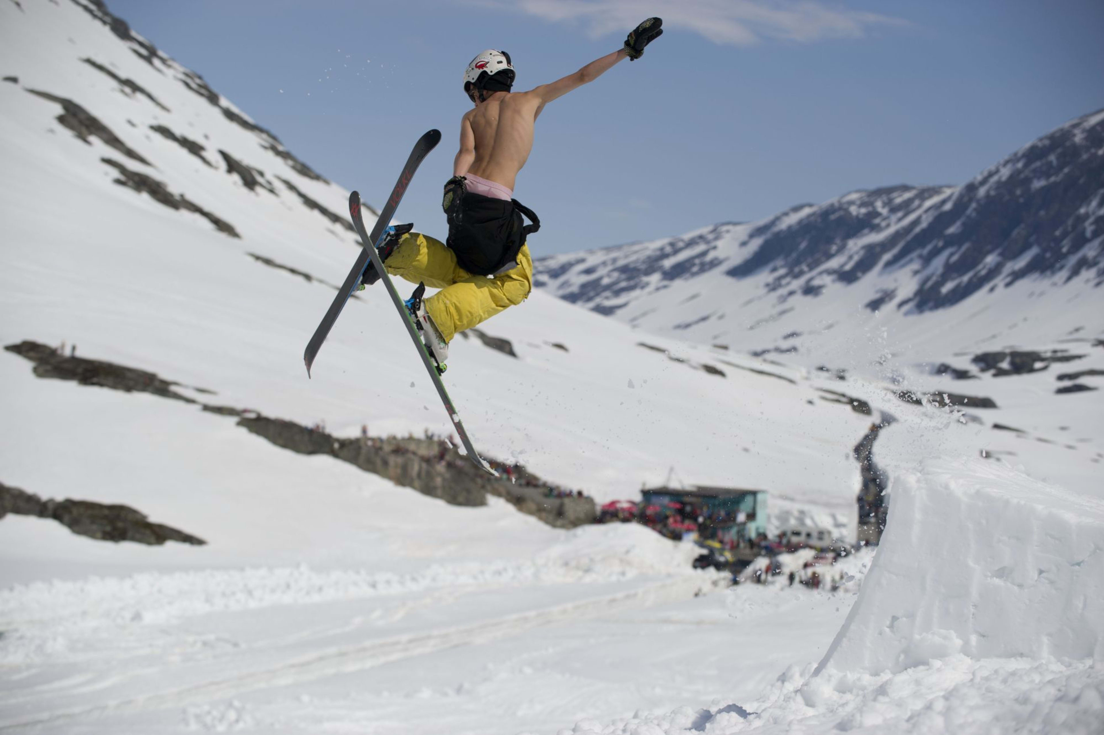 Ein Skifahrer in gelben Hosen führt einen Lufttrick von einem Schneehügel in einem Bergpark aus, mit einer jubelnden Menge im Hintergrund.