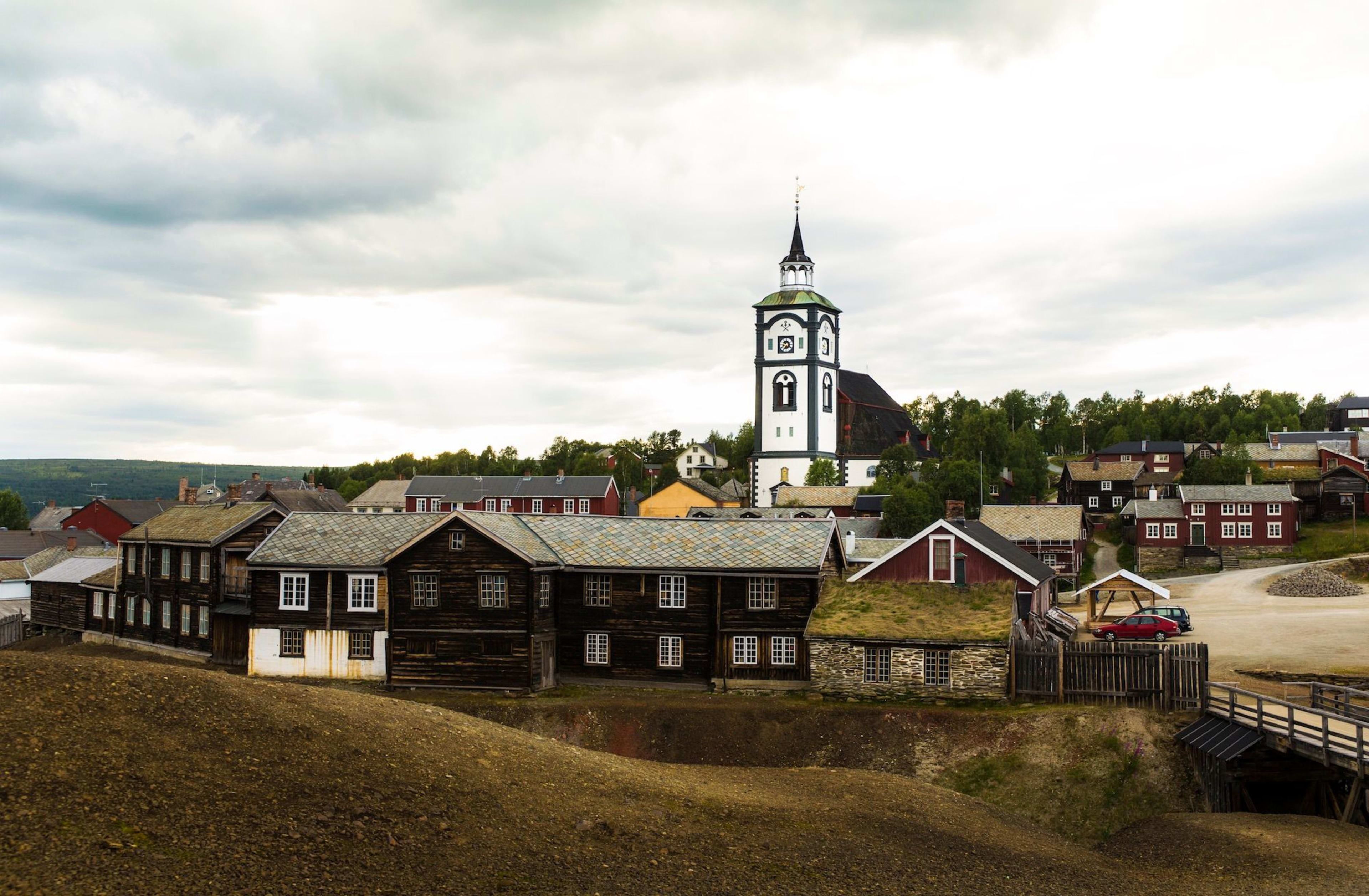 Historisk by med torvtak og mørke trehus, kirketårn i bakgrunnen.
