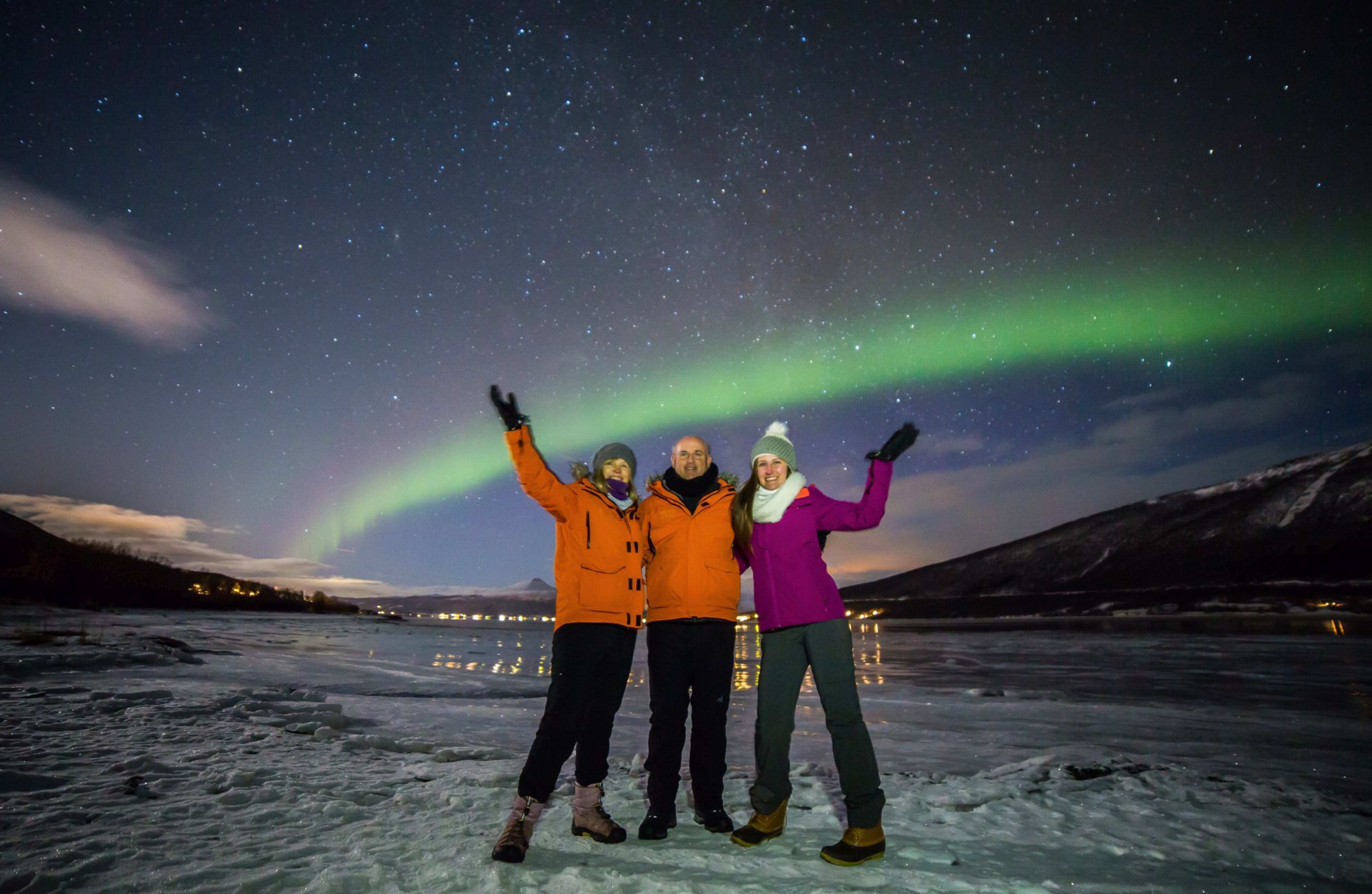 Fröhliche Gruppe von drei Personen in Winterkleidung unter grüner Aurora, Arme erhoben in Aufregung auf eisigem Gelände.