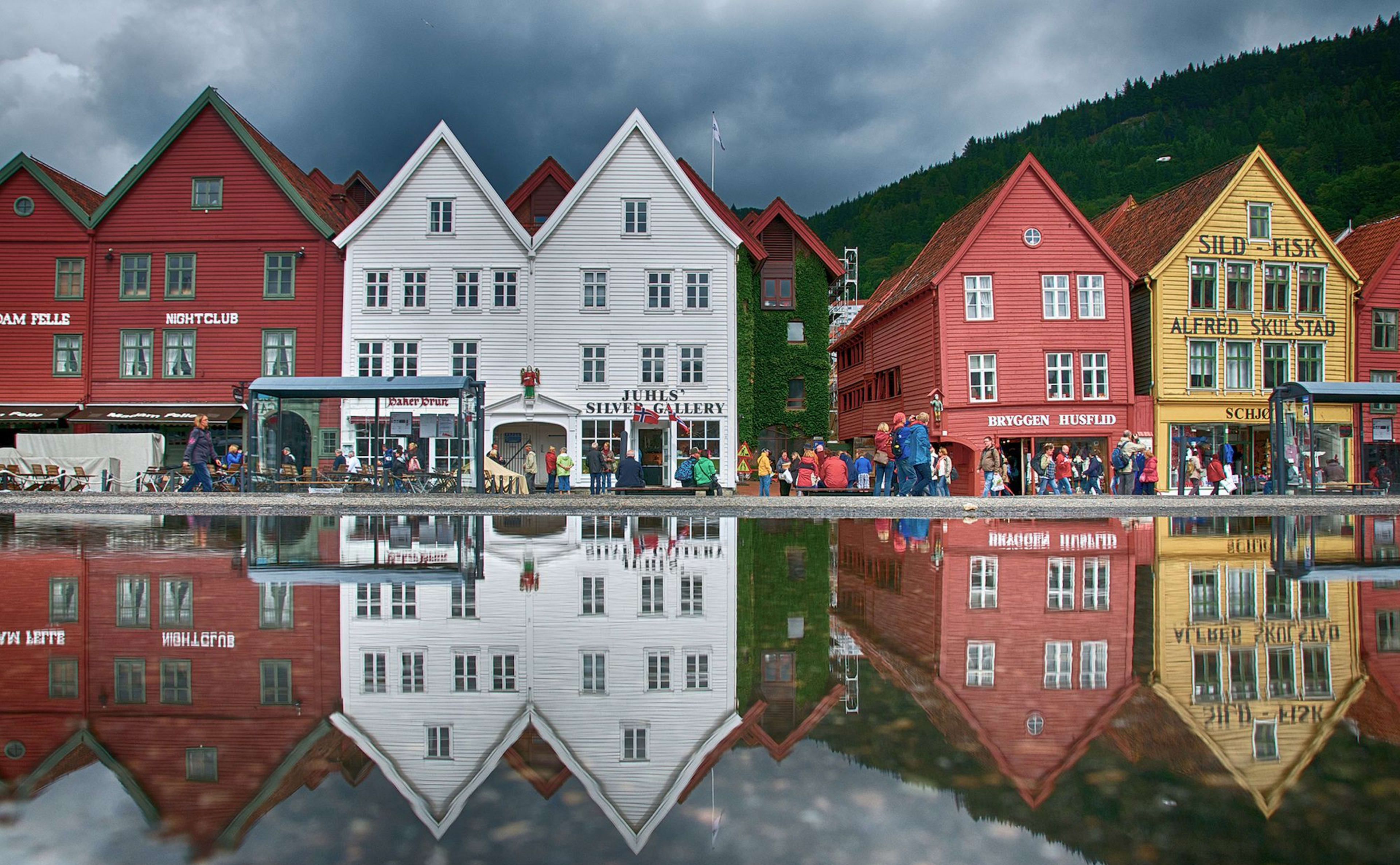 Fargerike Bryggen-bygninger i Bergen speiles i en hav, folk går under dramatiske skyer.