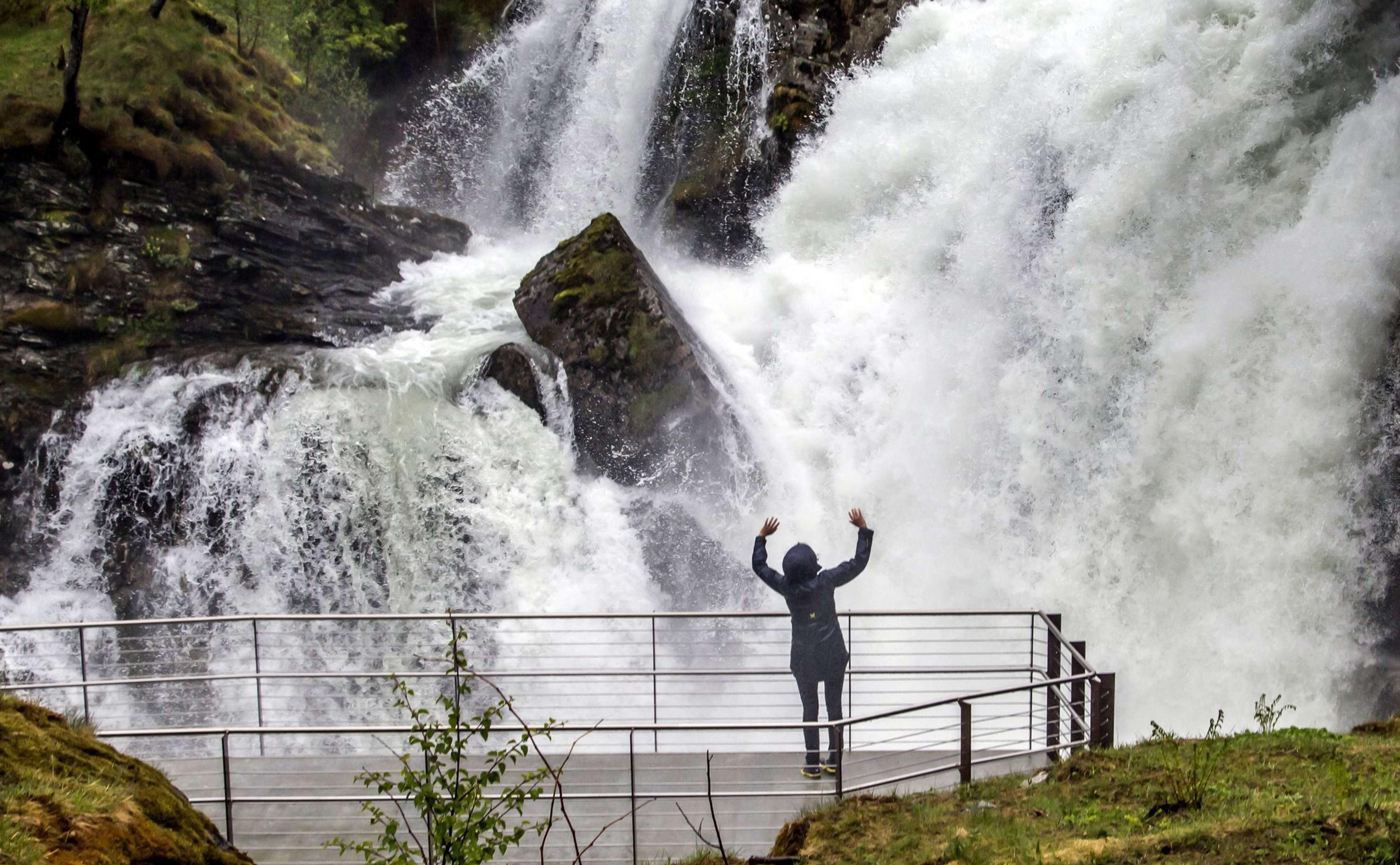 Person in rain jacket raises arms facing massive waterfall among greenery.