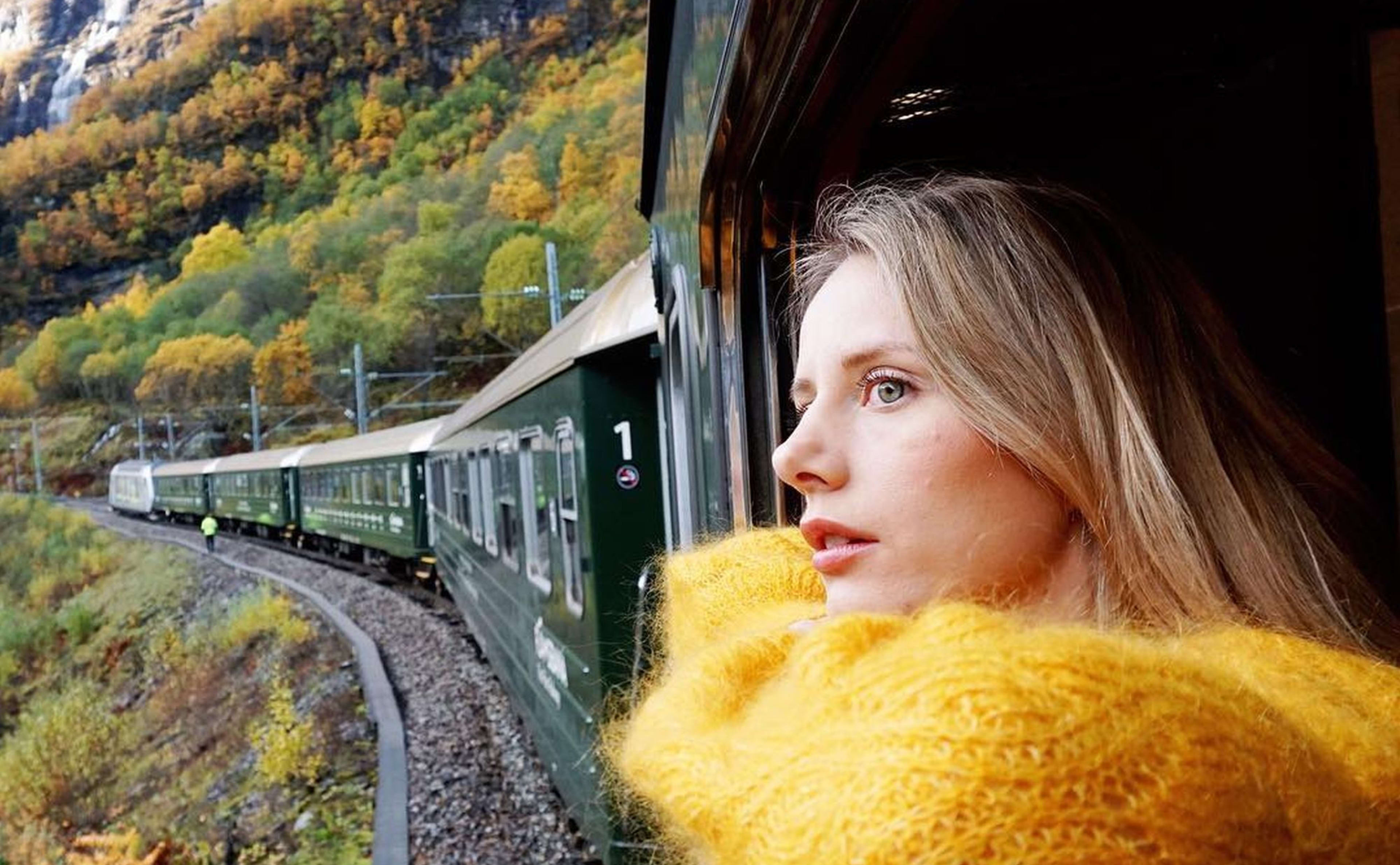 Frau im gelben Pullover blickt aus grünem Zugfenster auf herbstliche Landschaft.