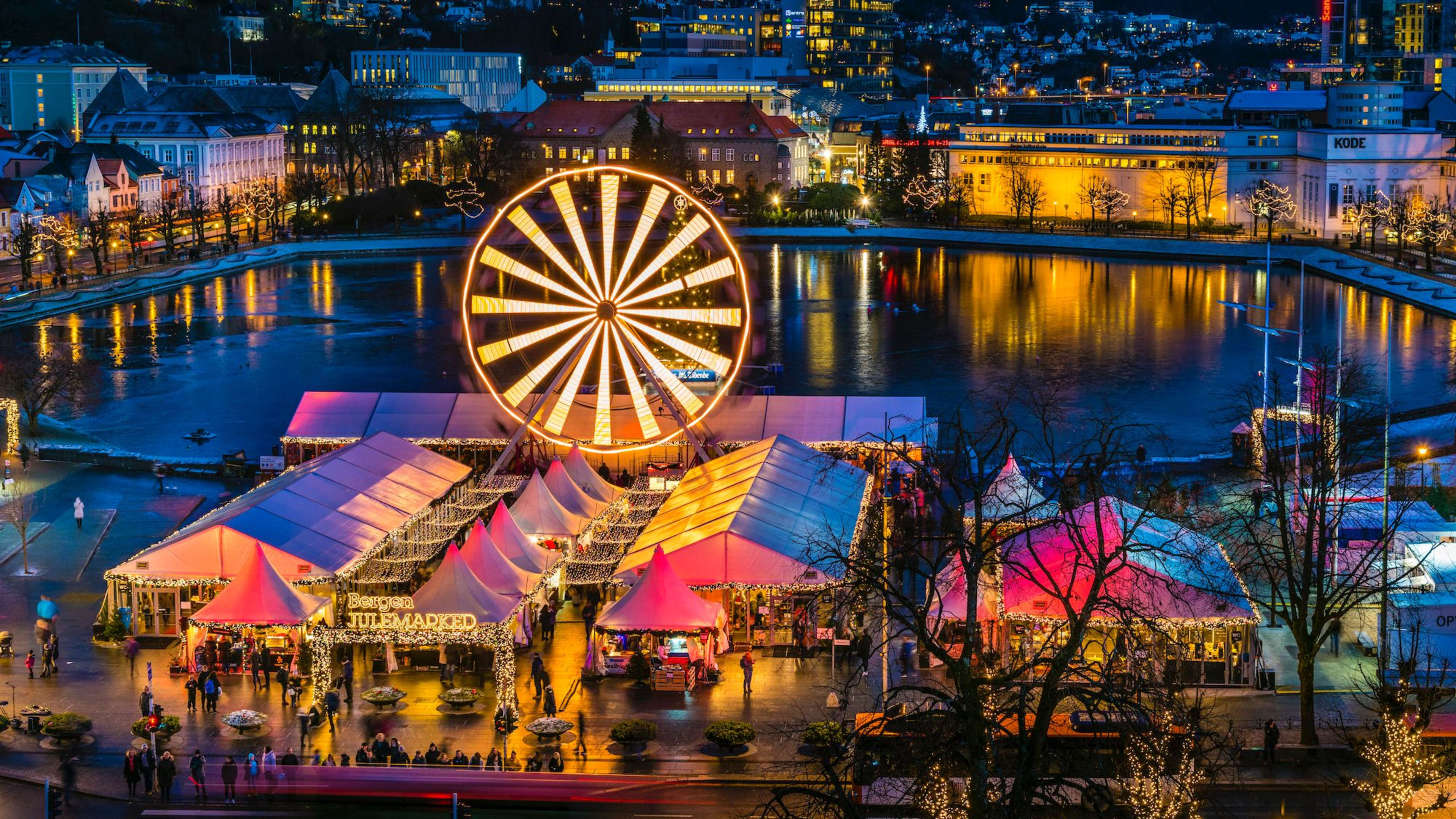 A Christmas market at night with a brightly lit Ferris wheel, festive tents, and a reflective body of water.