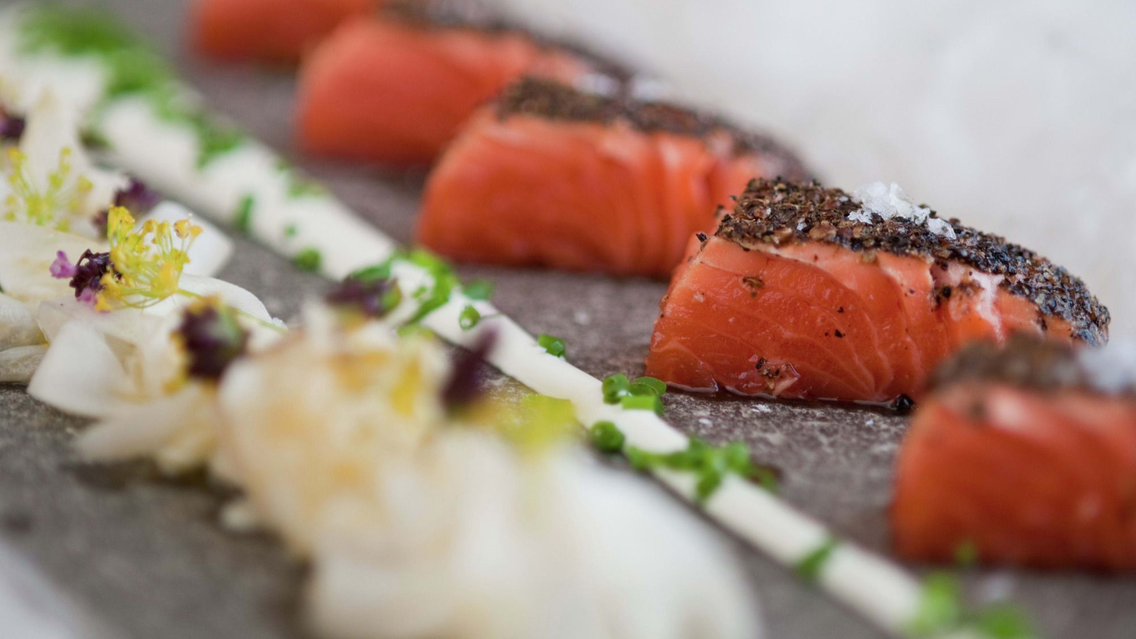 Eine elegante Präsentation von gepökeltem Lachs mit Pfefferkruste, cremiger Sauce und frischen Kräutern auf einem Stein.