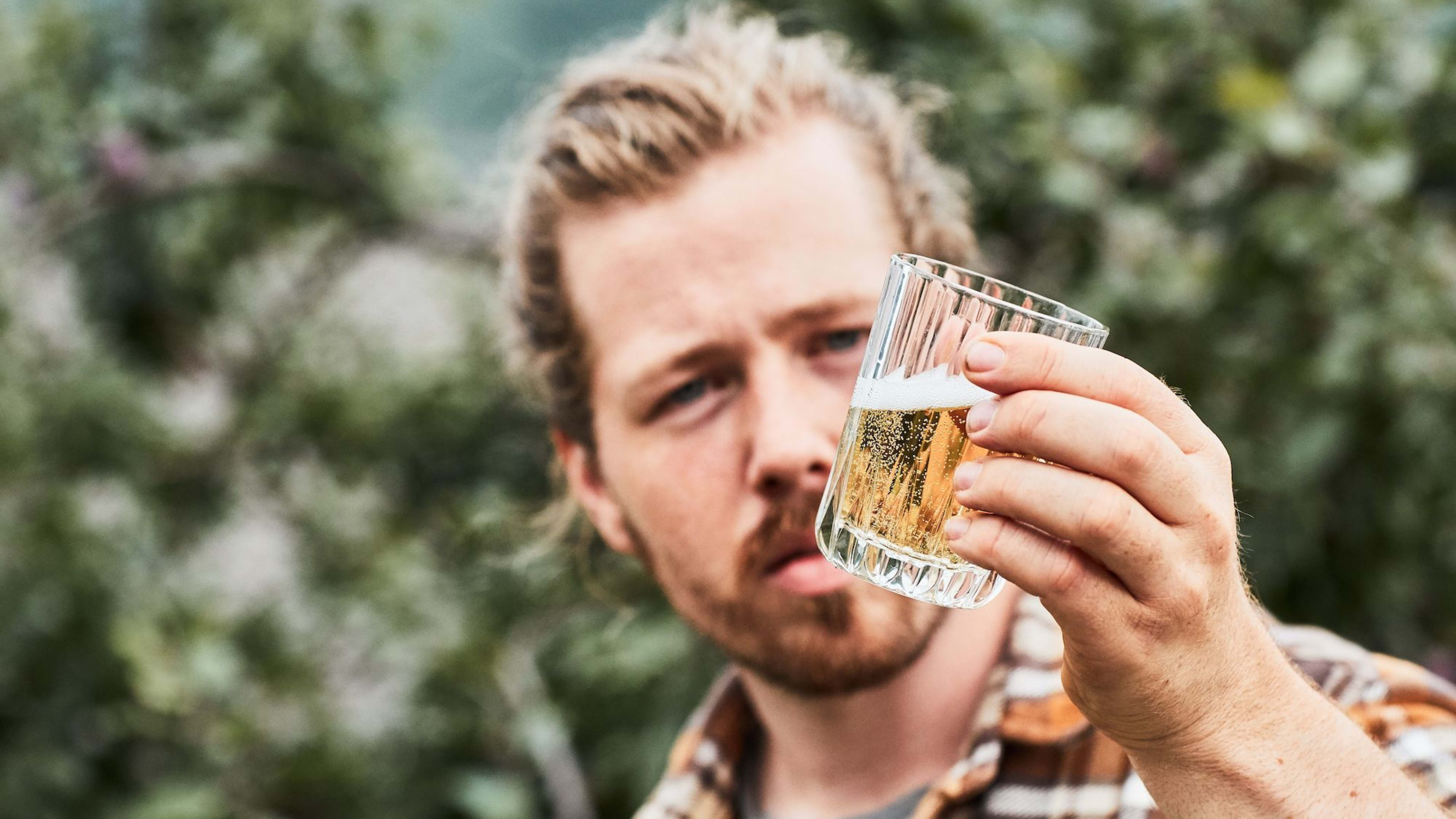 Mann im Karohemd betrachtet ein Glas Bier im Freien.