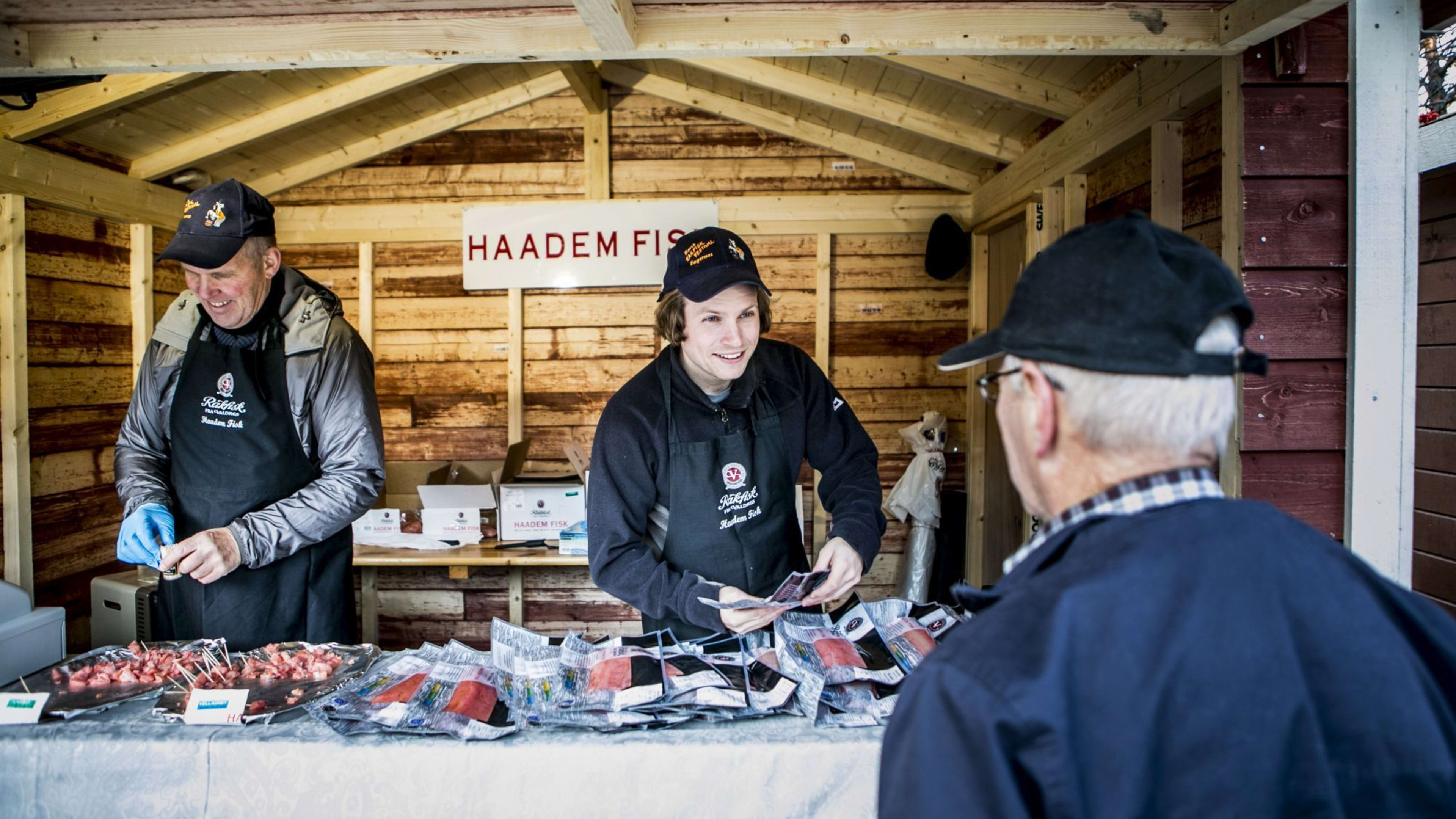 Markedsbod "Haadem Fish" med selgere som interagerer med kunder, rustikk treinteriør og fersk fisk til salgs.