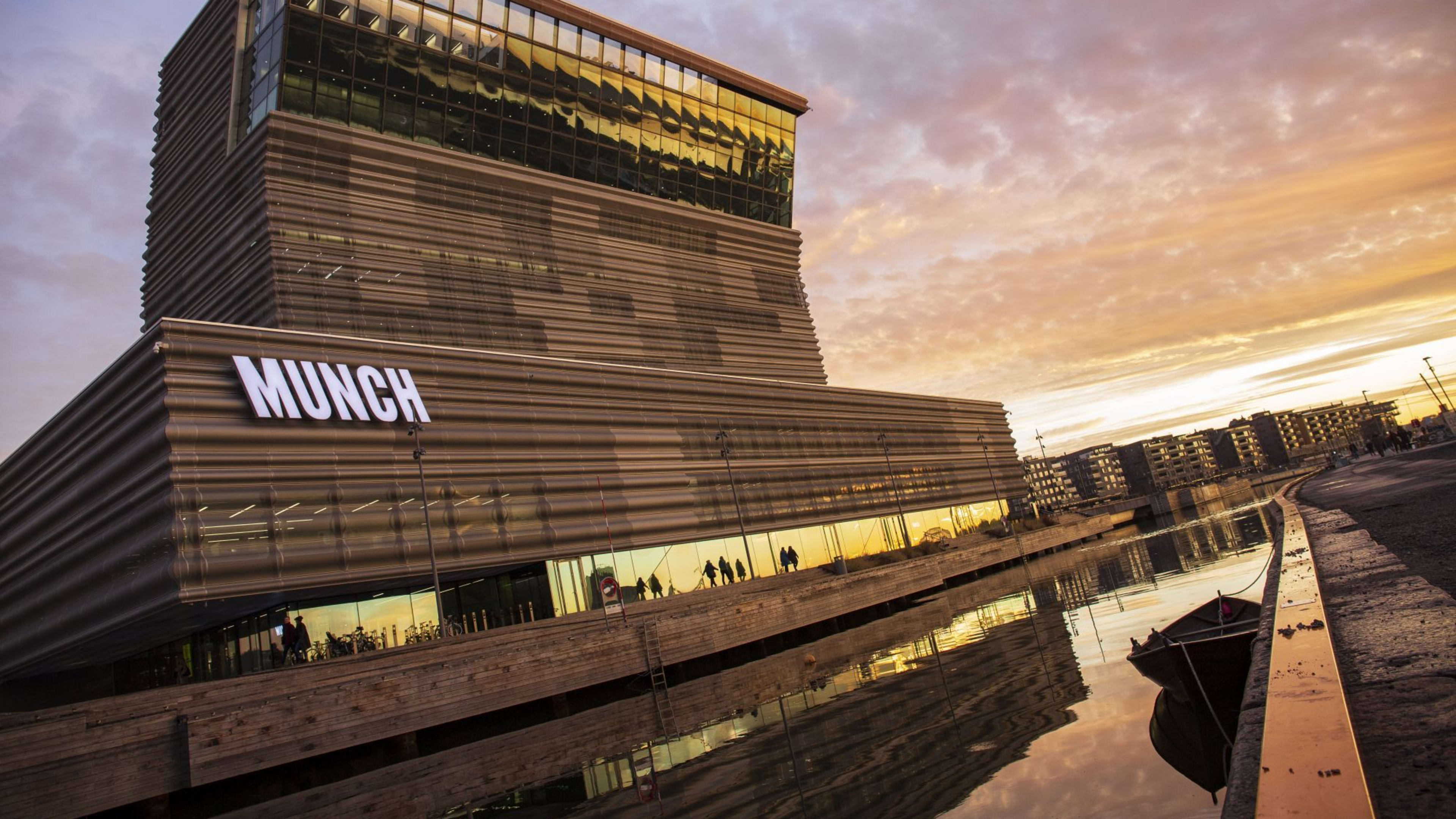 Munch-Museum mit modernem, kantigem Design, Glasfenstern und großem "MUNCH"-Schild, das sich im ruhigen Wasser bei Sonnenuntergang spiegelt.