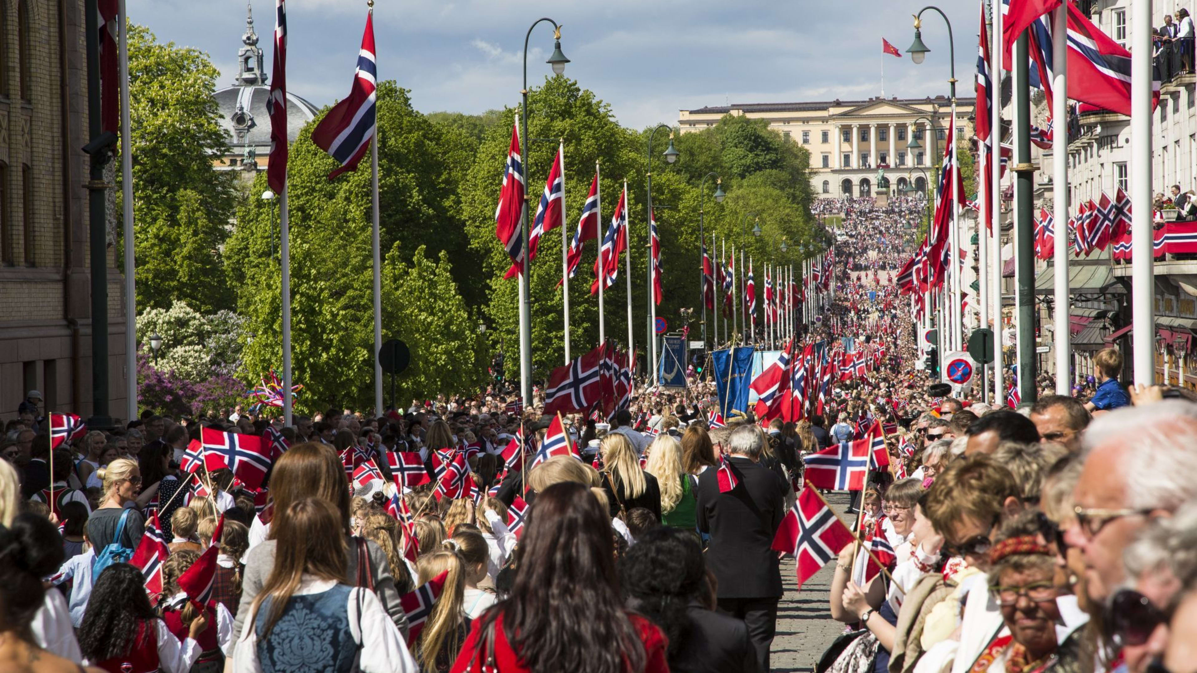 Menschenmenge feiert nationale Veranstaltung mit norwegischen Flaggen, traditionellen Kostümen und dem Königspalast im Hintergrund.