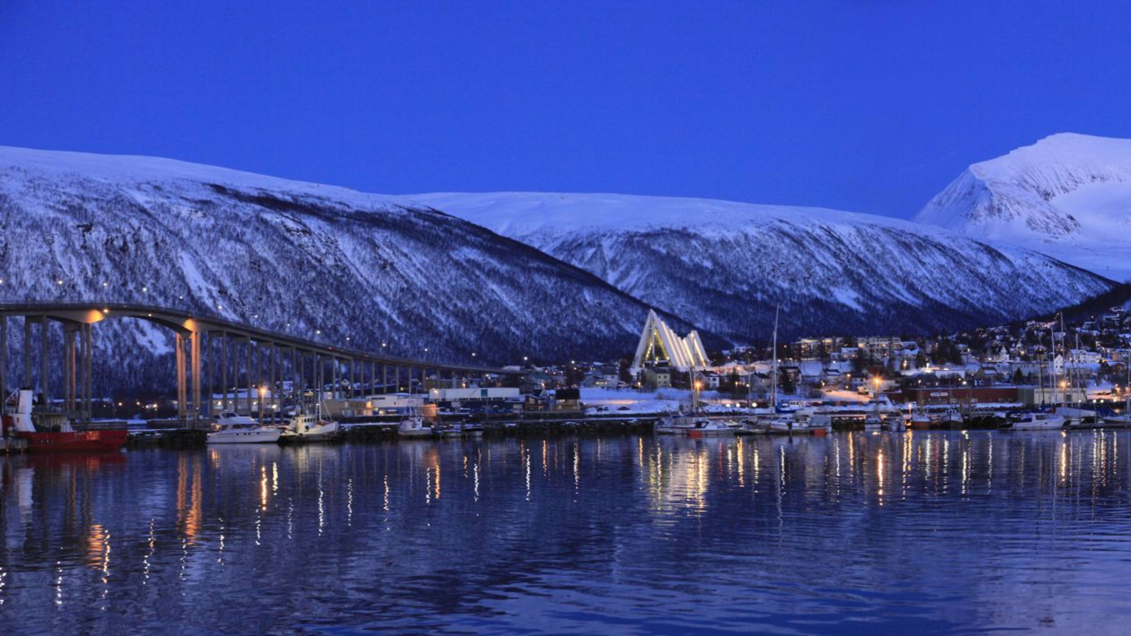 Panoramic winter view of Tromsø harbor with boats, snow-capped mountains, Arctic Cathedral, and reflected city lights.
