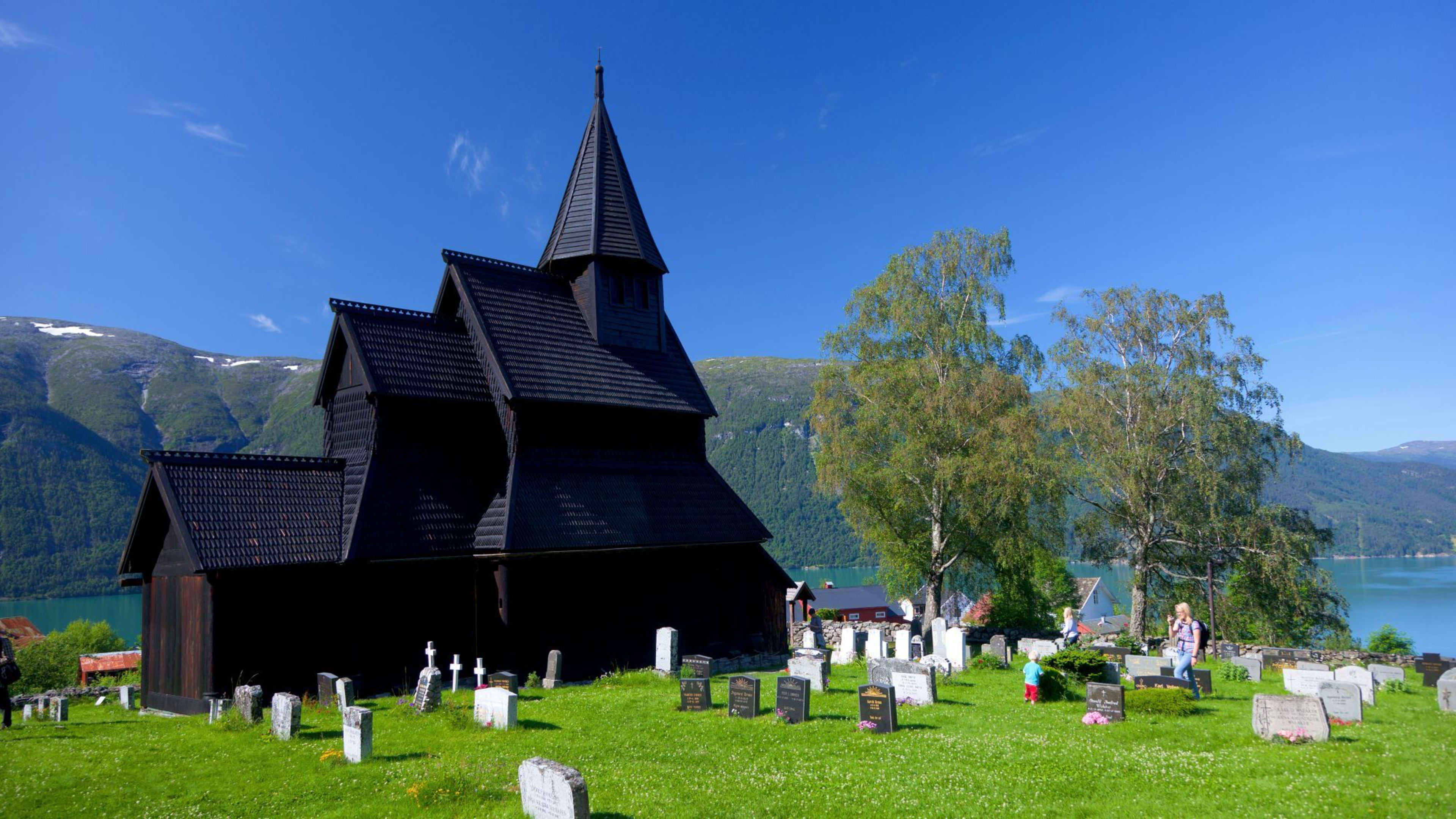 En svart trebent stavkirke med bratte tak står mot grønne bakker, en innsjø og en lys blå himmel.