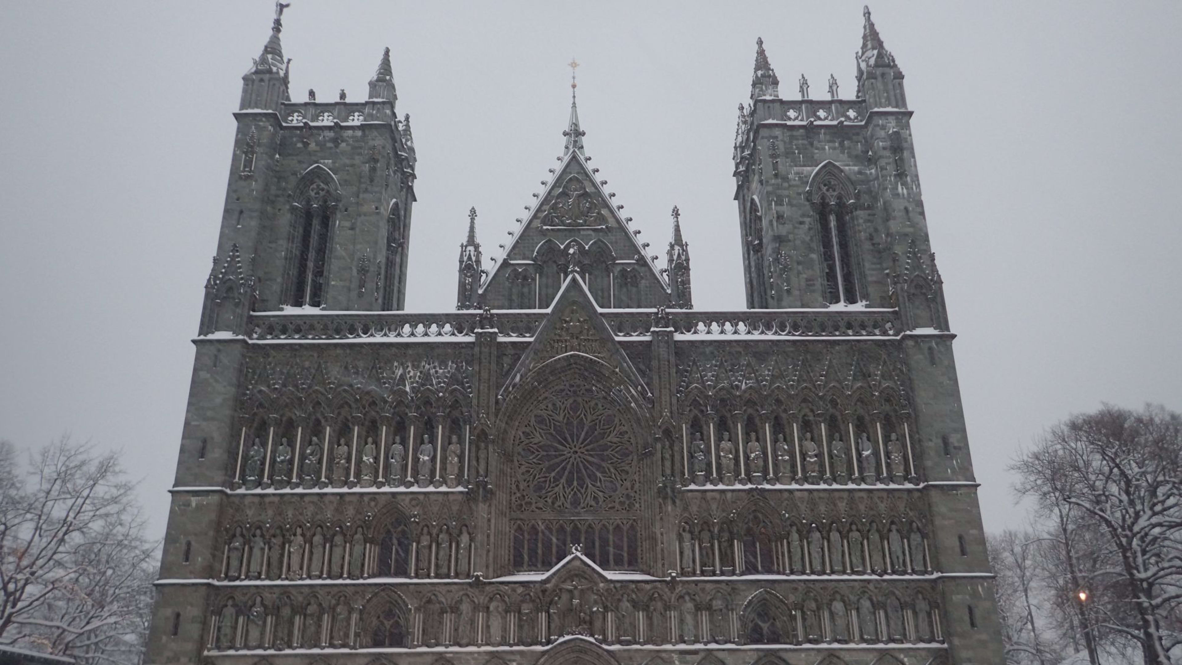 Gotische Kathedrale mit Schnee, kunstvollen Schnitzereien, Türmen und Rosenfenster, mit Menschen im verschneiten Innenhof unter grauem Himmel.