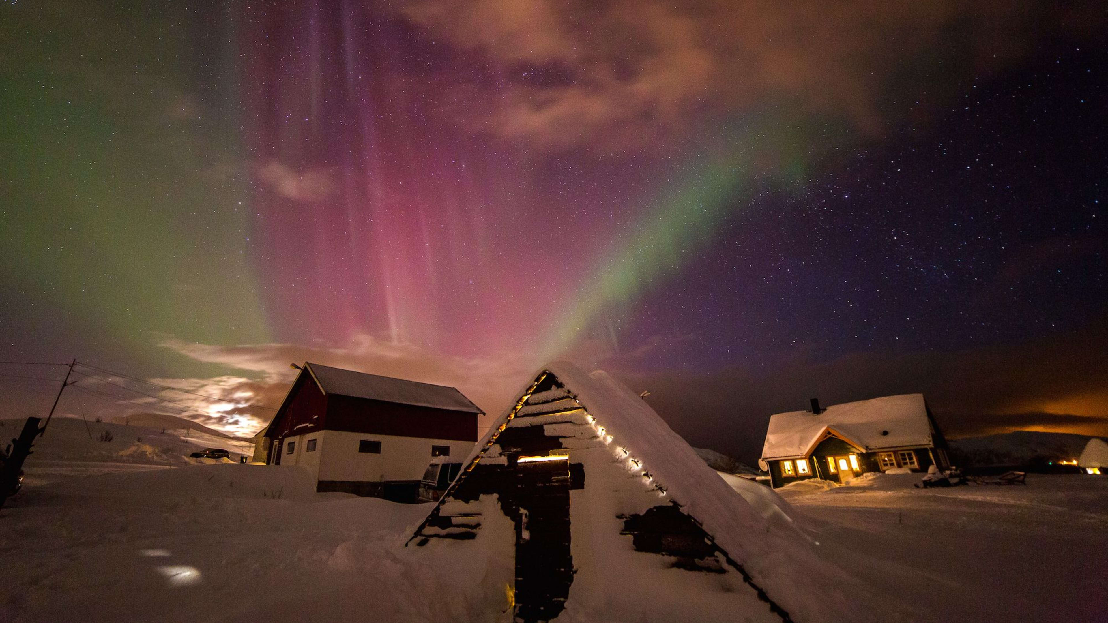 Nordlys i grønne og rosa nyanser over snødekte hus, koselige hytter med varmt lys som skaper et magisk vintersceneri.