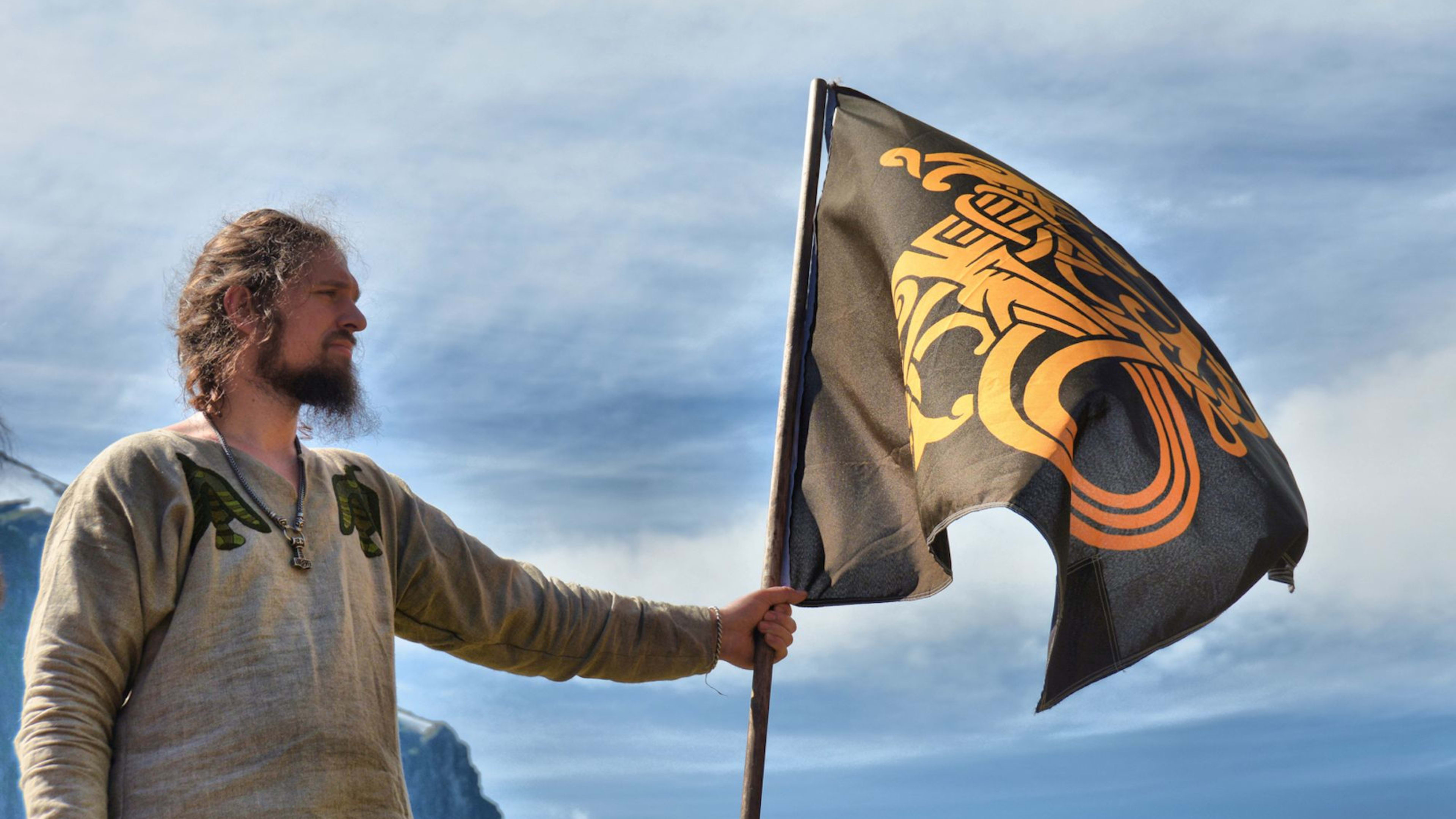 Mann i vikinginspirert klær holder svart flagg med nordisk kunst, dramatiske fjelltopper i bakgrunnen og skyet himmel.