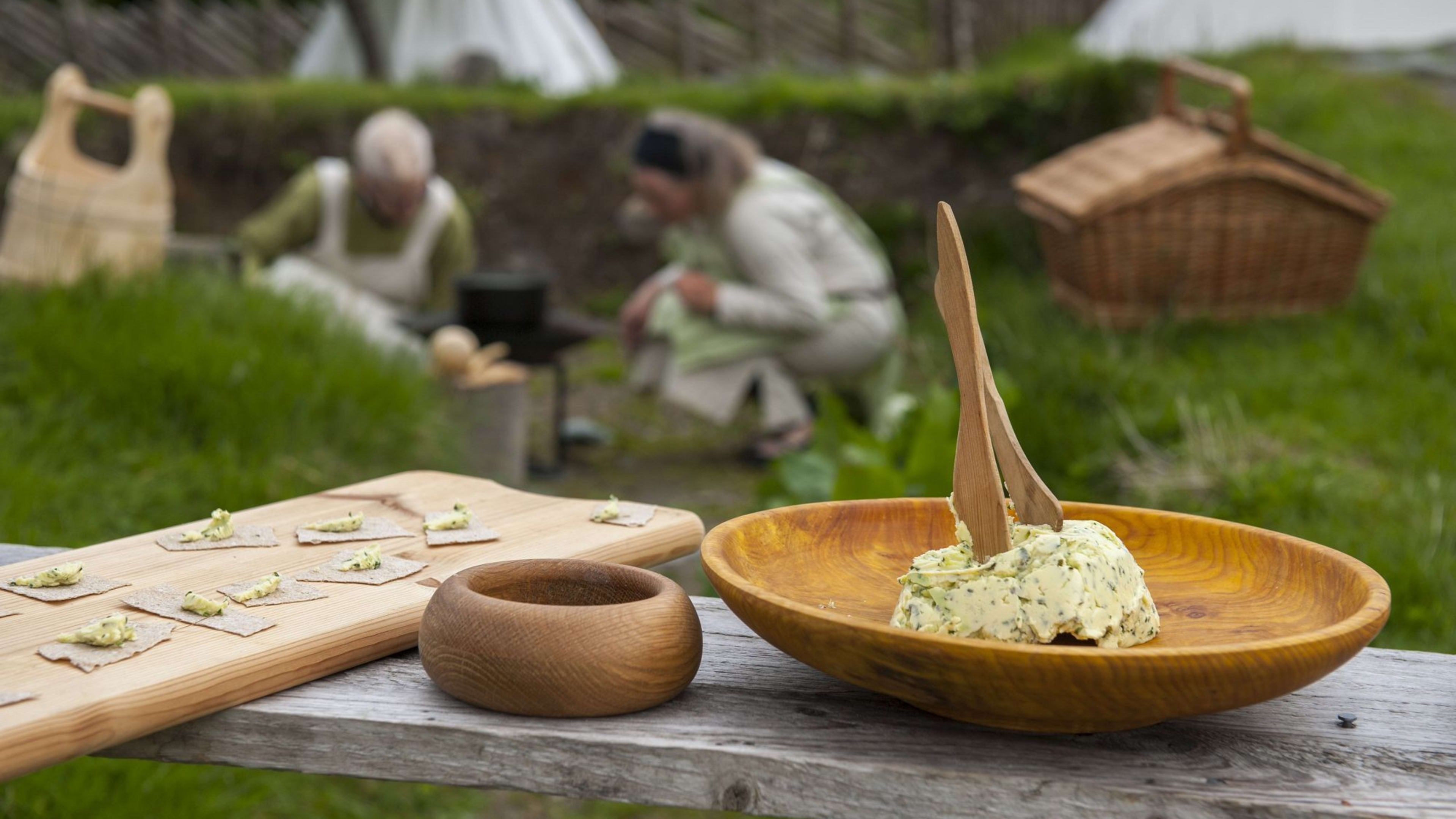Tre Vikingbord med matforberedelse, urtesmør, treboller og kjeks, med historiske figurer i bakgrunnen.
