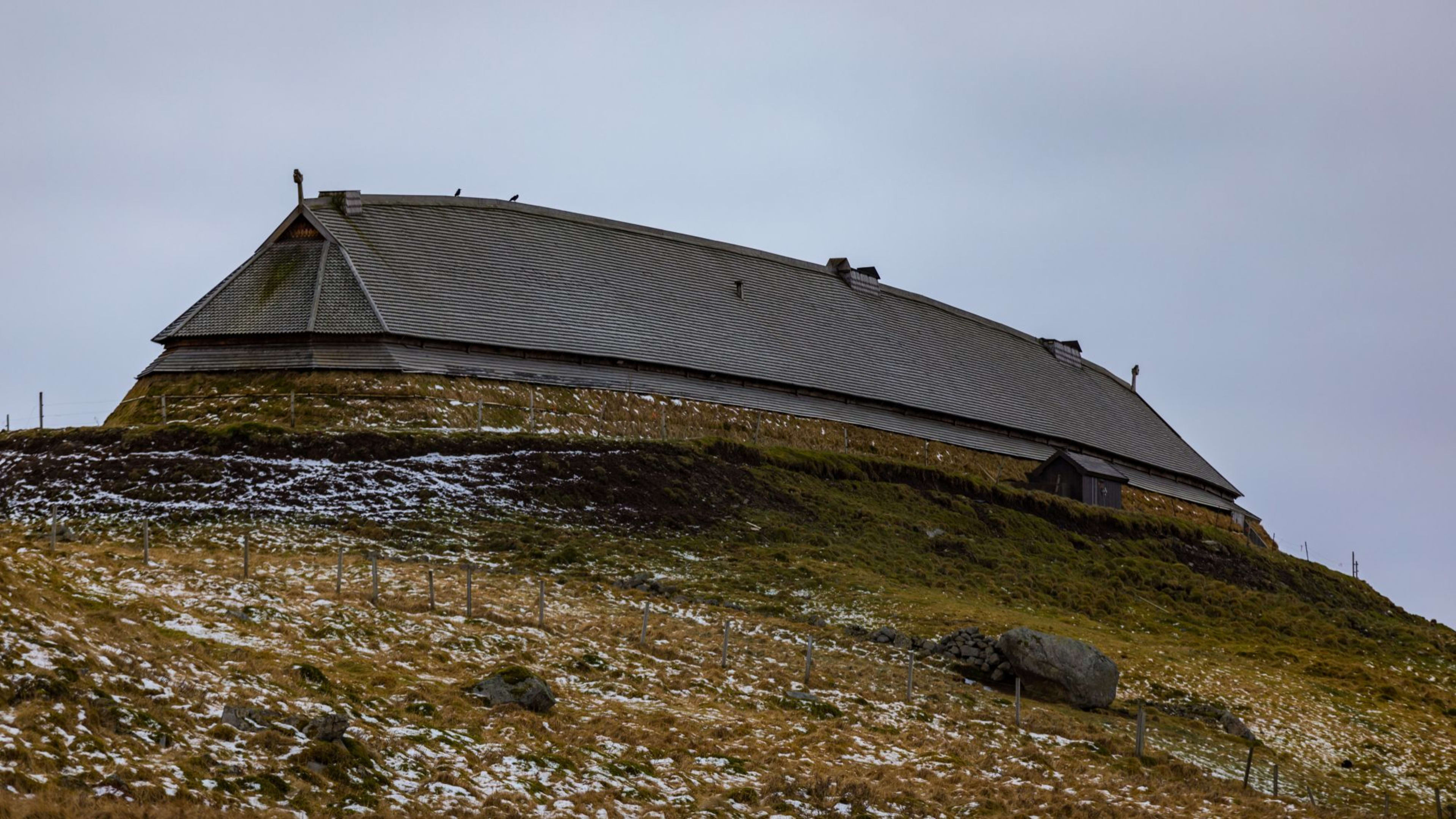 Wikinger-Langhaus auf einem grasbewachsenen Hügel mit Schneeflecken, Holzstruktur, bewölkter Himmel, der eine düstere, kalte Atmosphäre schafft.