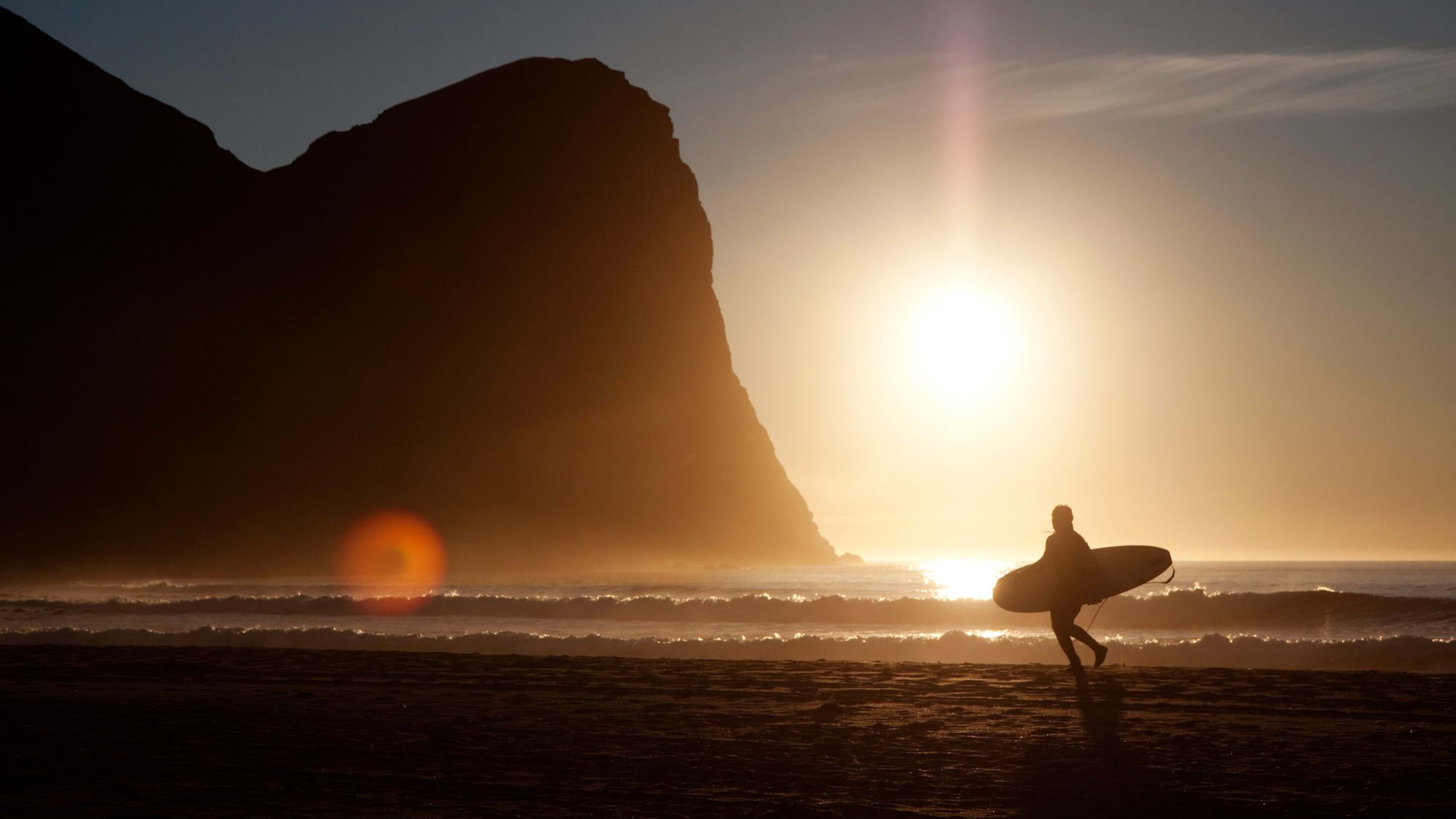 Eine Silhouette eines Surfers, der am Strand bei Sonnenuntergang entlang geht und ein Surfbrett trägt.