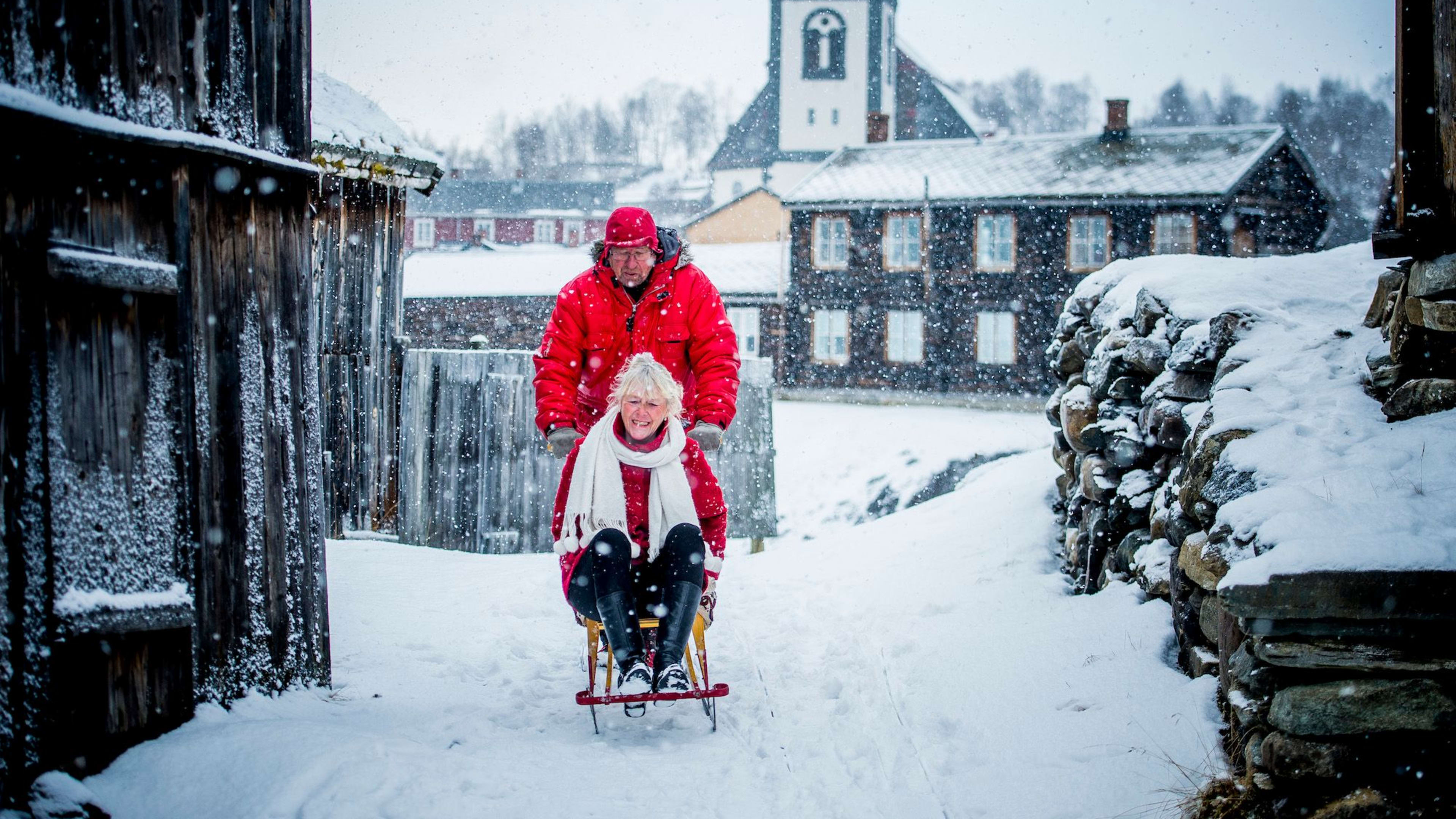 Älteres Paar genießt Schnee, Frau lacht auf Schlitten, Mann schiebt.