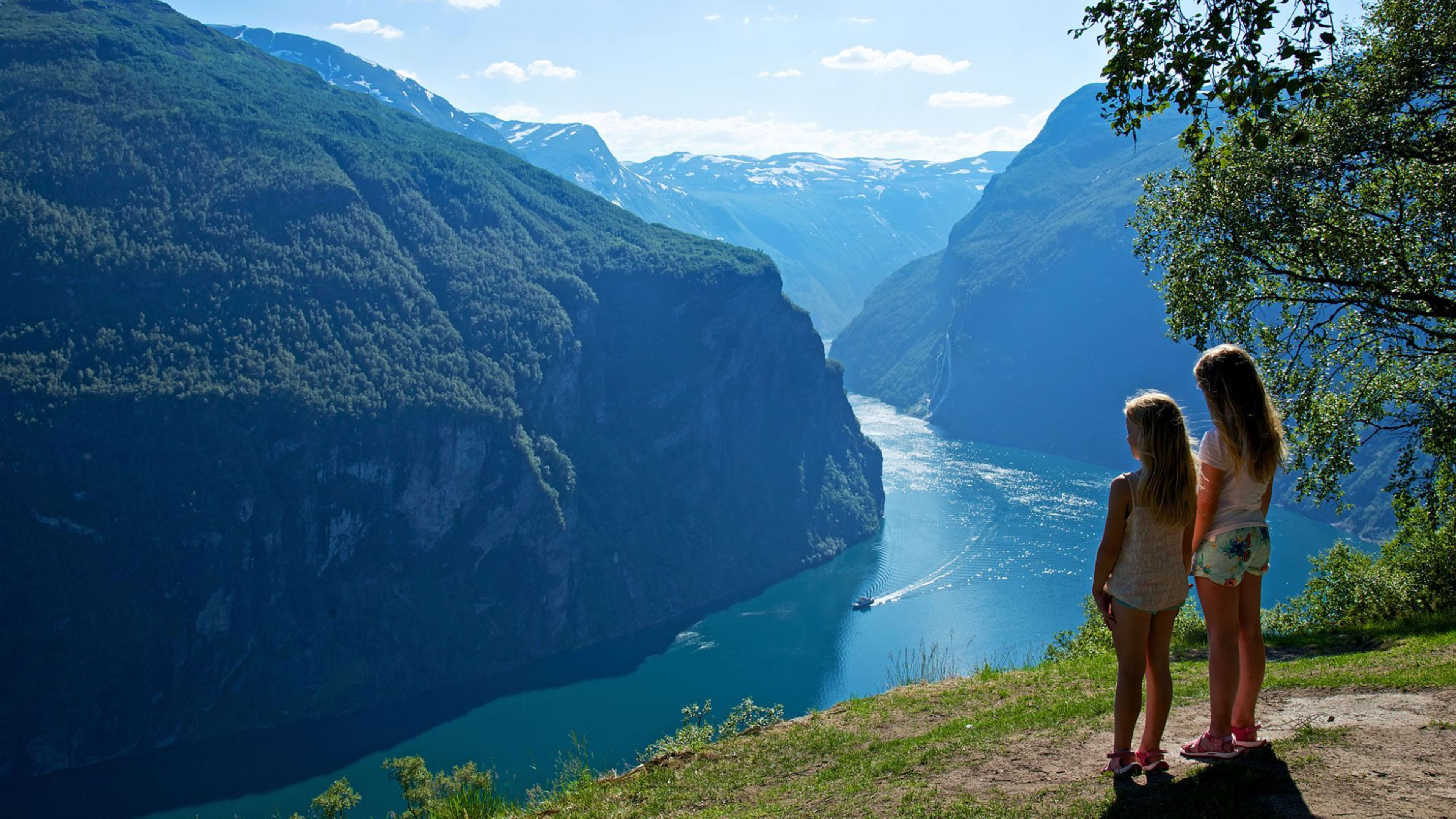 To jenter ser på norsk fjord, klipper dekket med grønt, rolig blått vann som reflekterer den lyse himmelen.